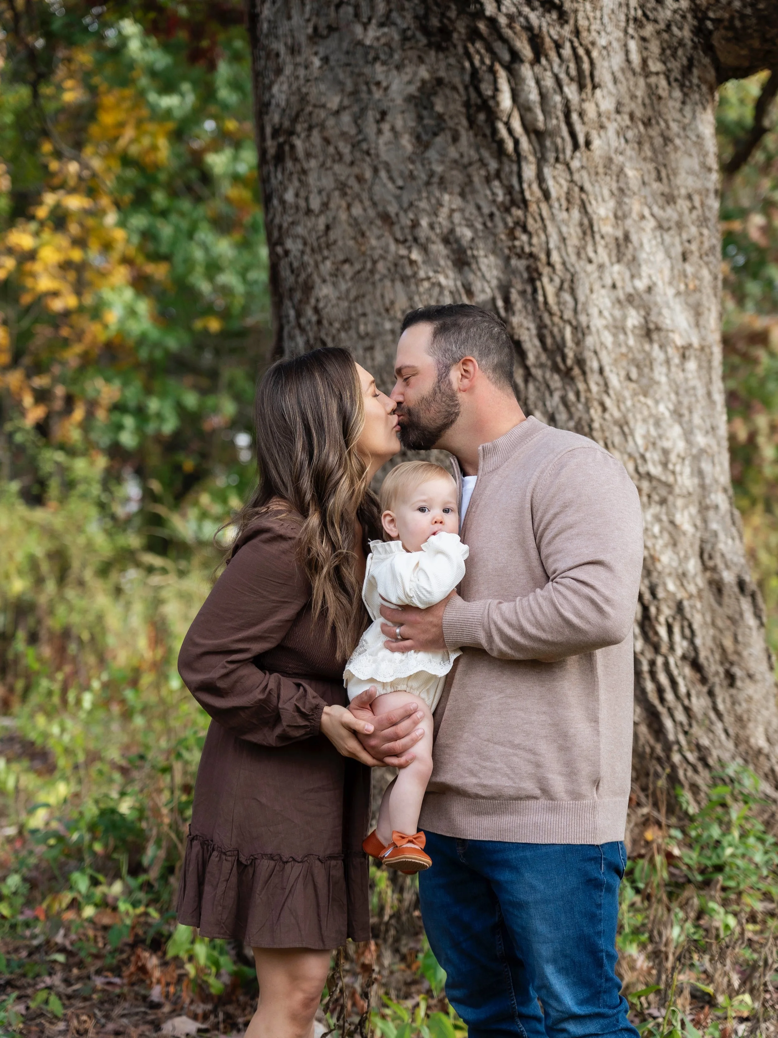 A family of three standing outdoors near a large tree, with the parents about to kiss and the baby child being held by the father.