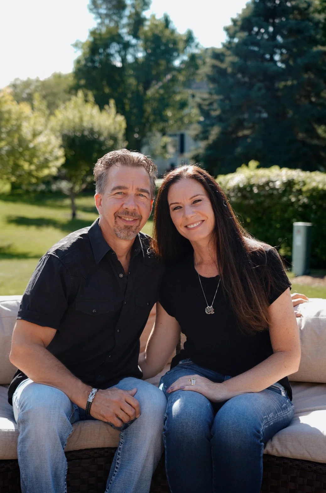A smiling man and woman sitting closely together on an outdoor cushioned sofa in a park with green grass and trees in the background, enjoying a sunny day.