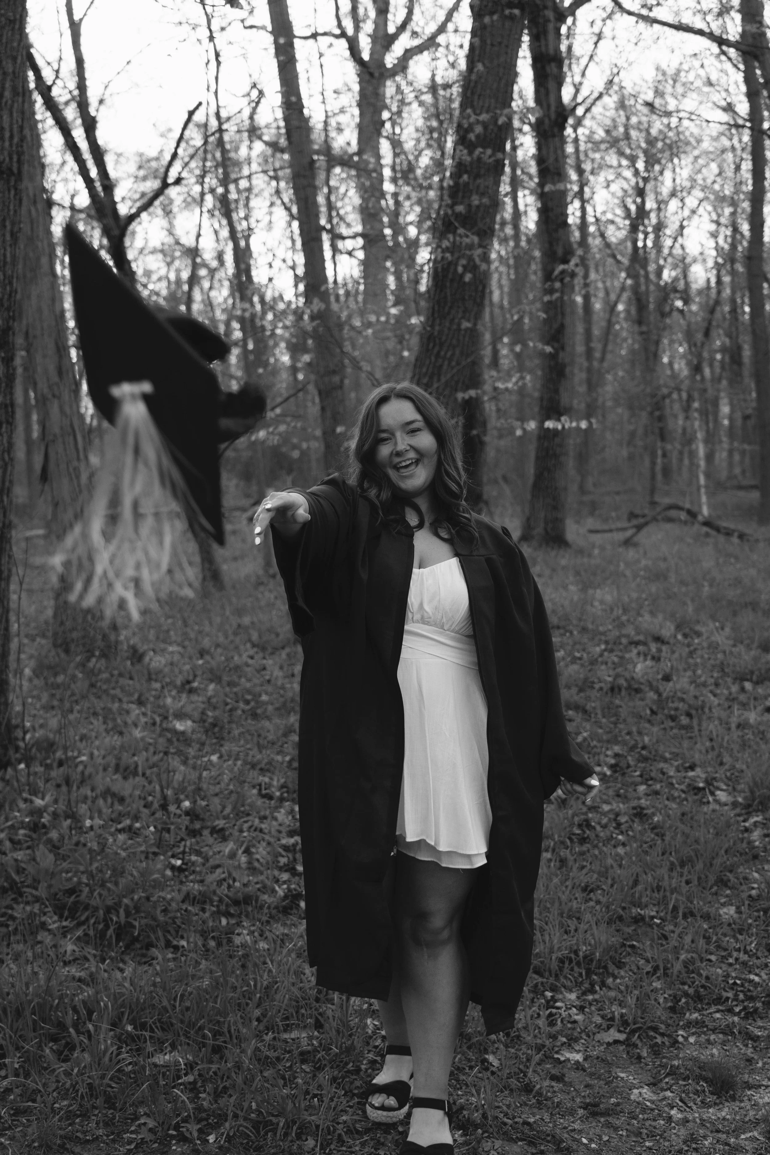 A young woman in a graduation gown and cap celebrating in a wooded area, smiling, with her cap in the foreground.