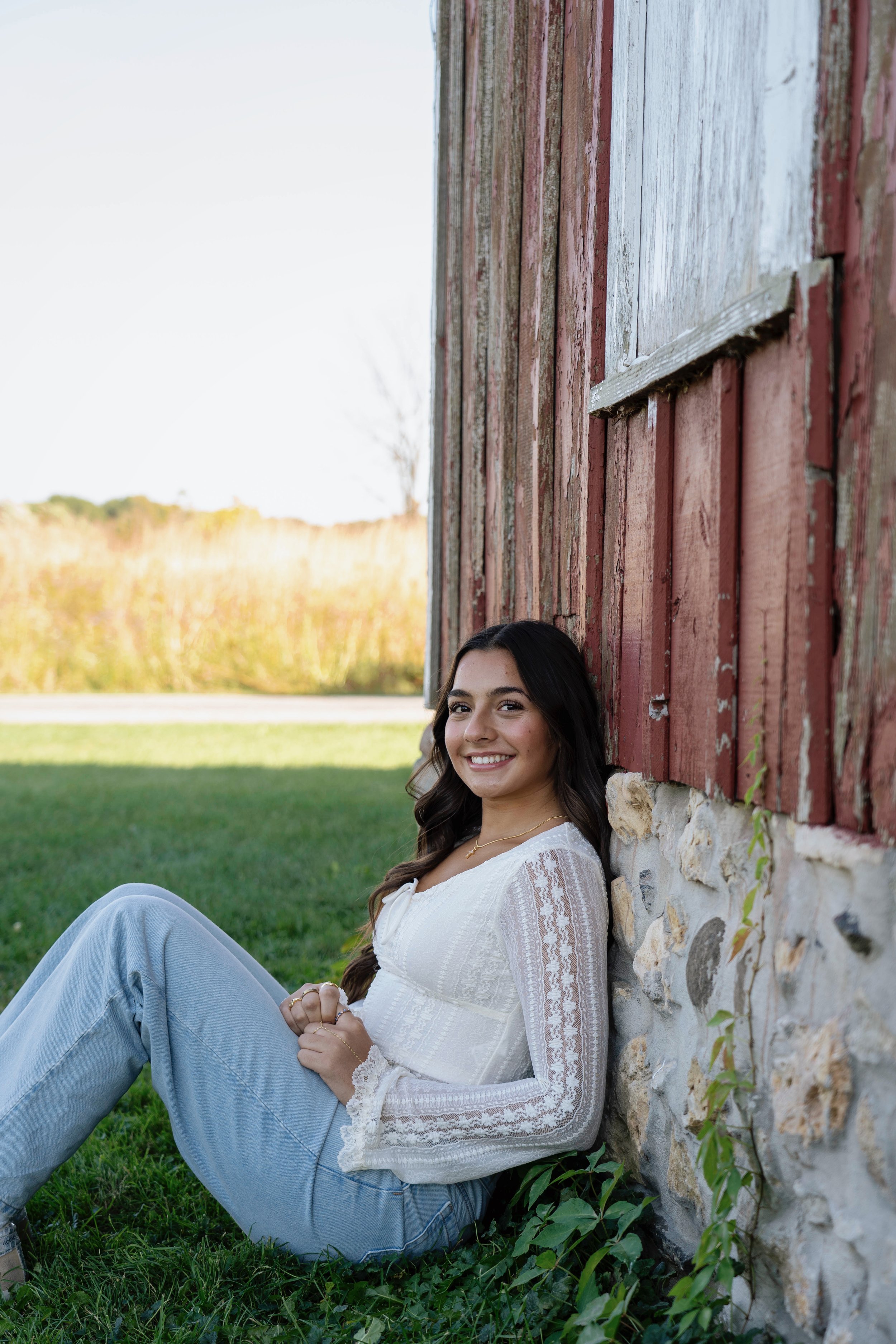 A young woman with dark hair and a white lace blouse sitting on green grass next to a rustic red wooden building, smiling at the camera.