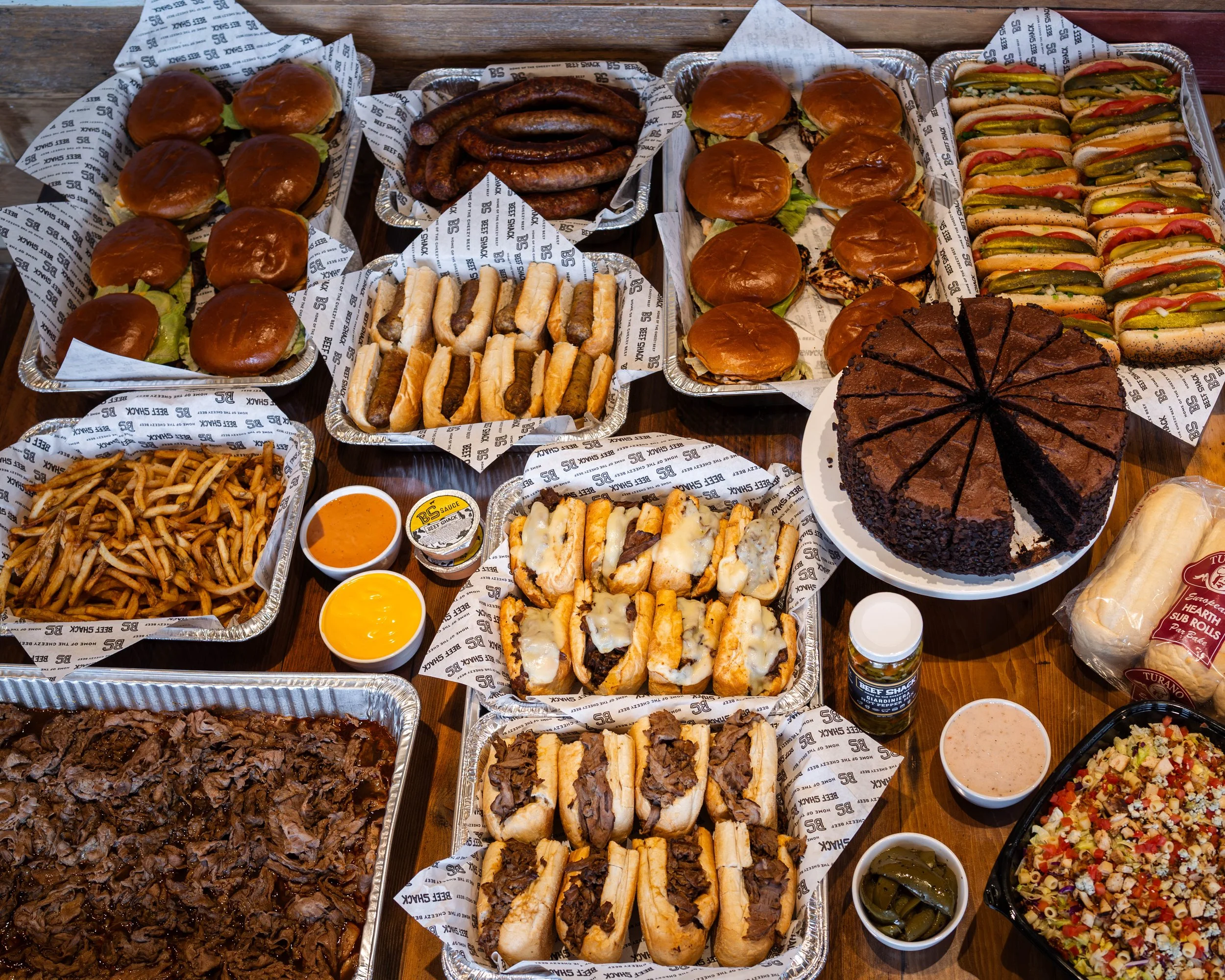 Spread of barbecue food including sliders, hot dogs, sausage links, fries, a chocolate cake, baked beans, salad, dipping sauces, and rolls on a wooden table.