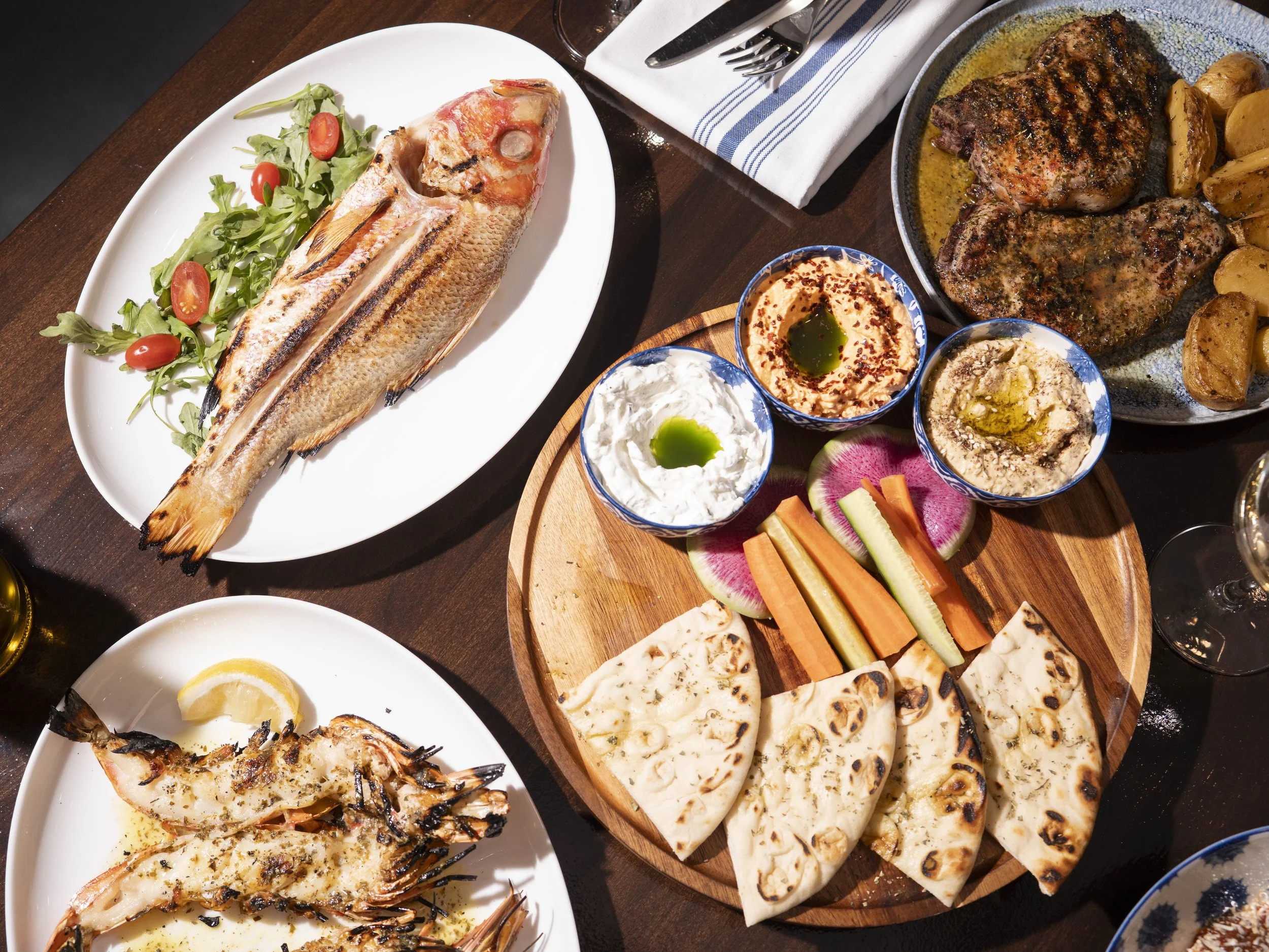 A variety of Middle Eastern dishes including grilled fish with salad, grilled meat with potatoes, roasted shrimp with lemon, and assorted dips with vegetables and flatbread on a wooden platter.