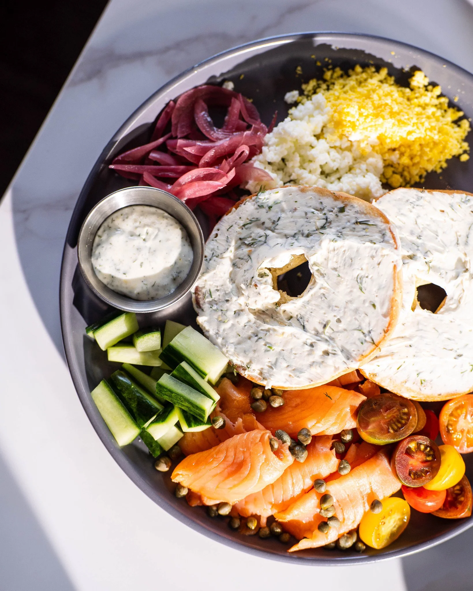 A platter with smoked salmon, cherry tomatoes, cucumber slices, creamy herb cheese, pickled onions, cauliflower, grated cheese, dill, capers, and a small container of creamy herb sauce.