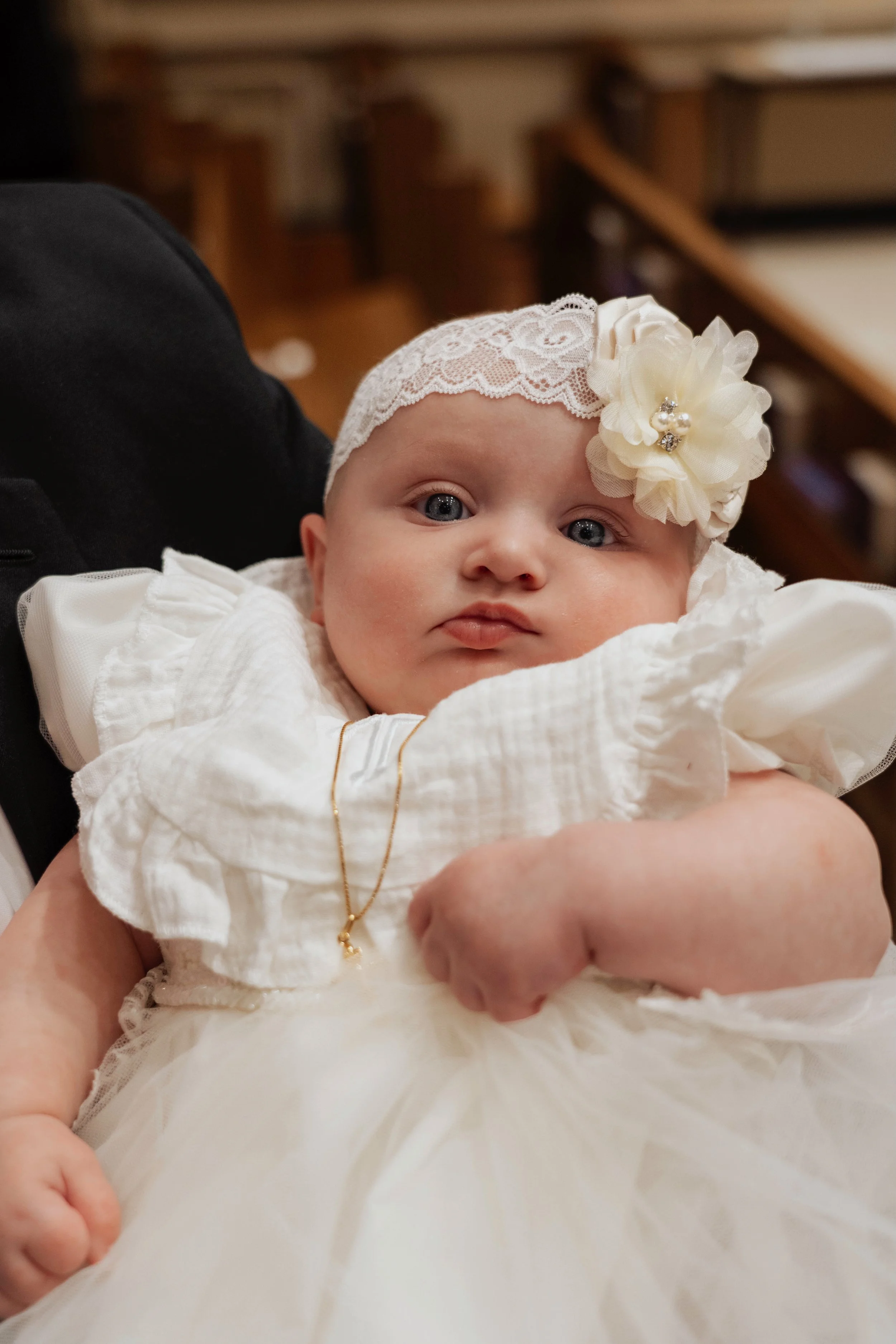 A baby girl dressed in a white lace headband with a flower and diamond accent, wearing a white dress with puffed sleeves and a gold necklace, is being held. The background suggests a church or formal setting.