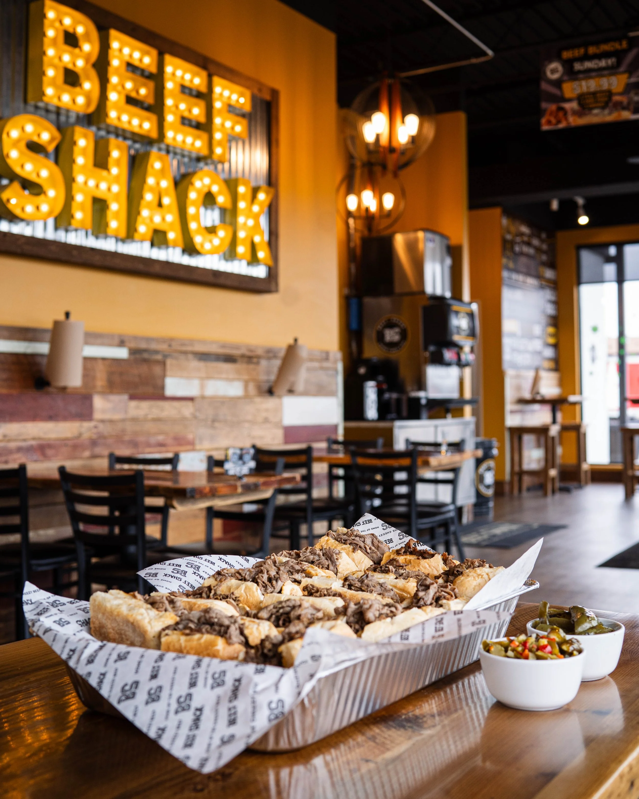 A basket of loaded hot dogs topped with shredded beef on a paper liner, accompanied by bowls of pickles and sliced peppers, inside a restaurant with a sign reading 'BEEF SHACK' illuminated with yellow lights.