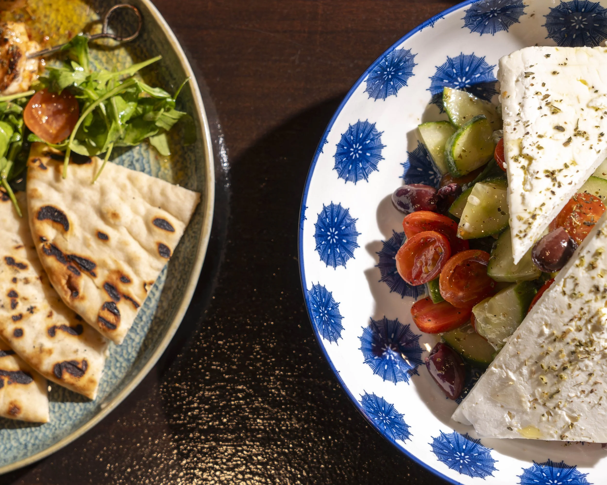 A plate of Greek salad with feta cheese, cherry tomatoes, cucumber, olives, and herbs, alongside a dish of grilled pita bread with salad.