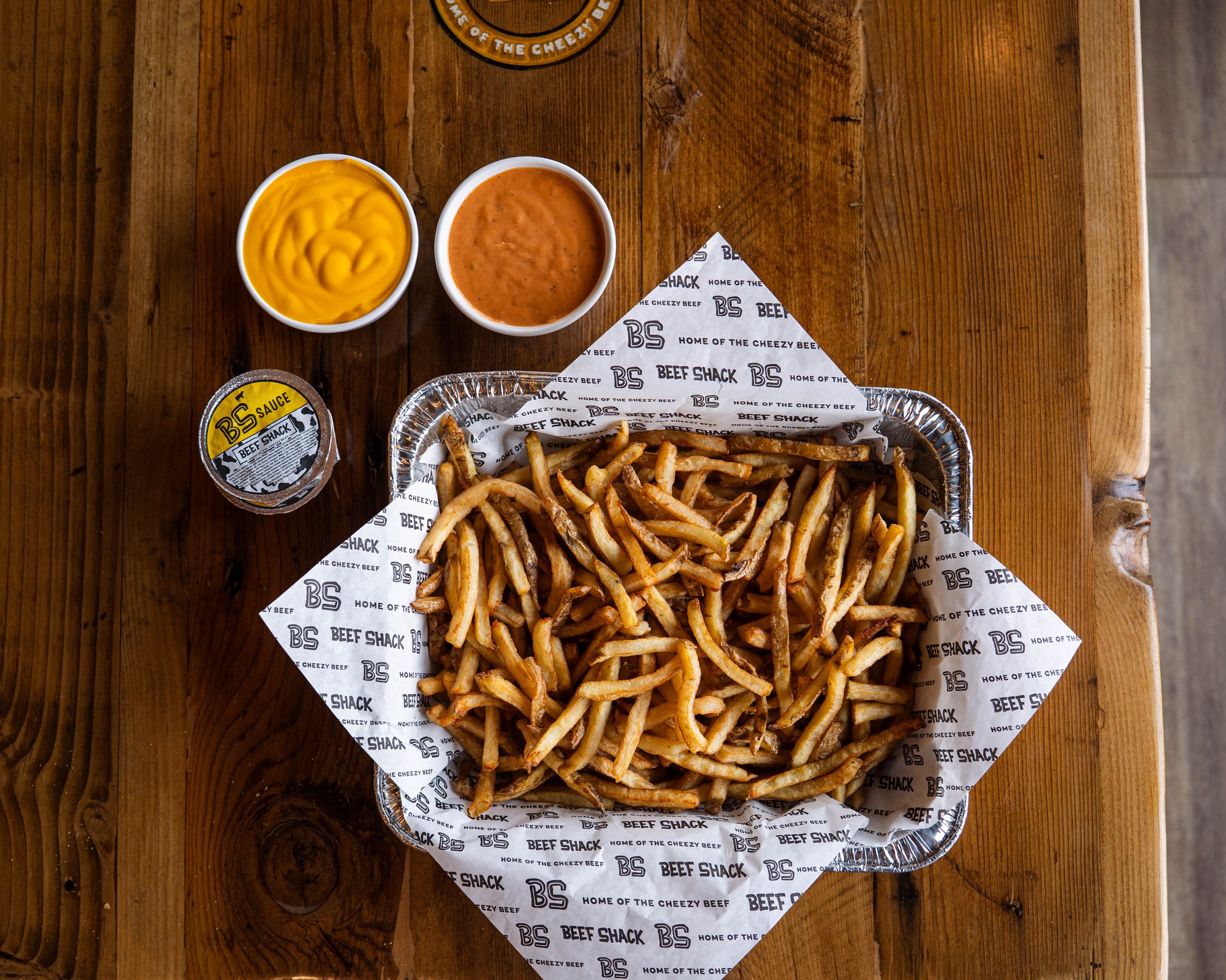 Basket of French fries on branded paper, with containers of mustard and barbecue sauce, and a container of dipping sauce on a wooden table.