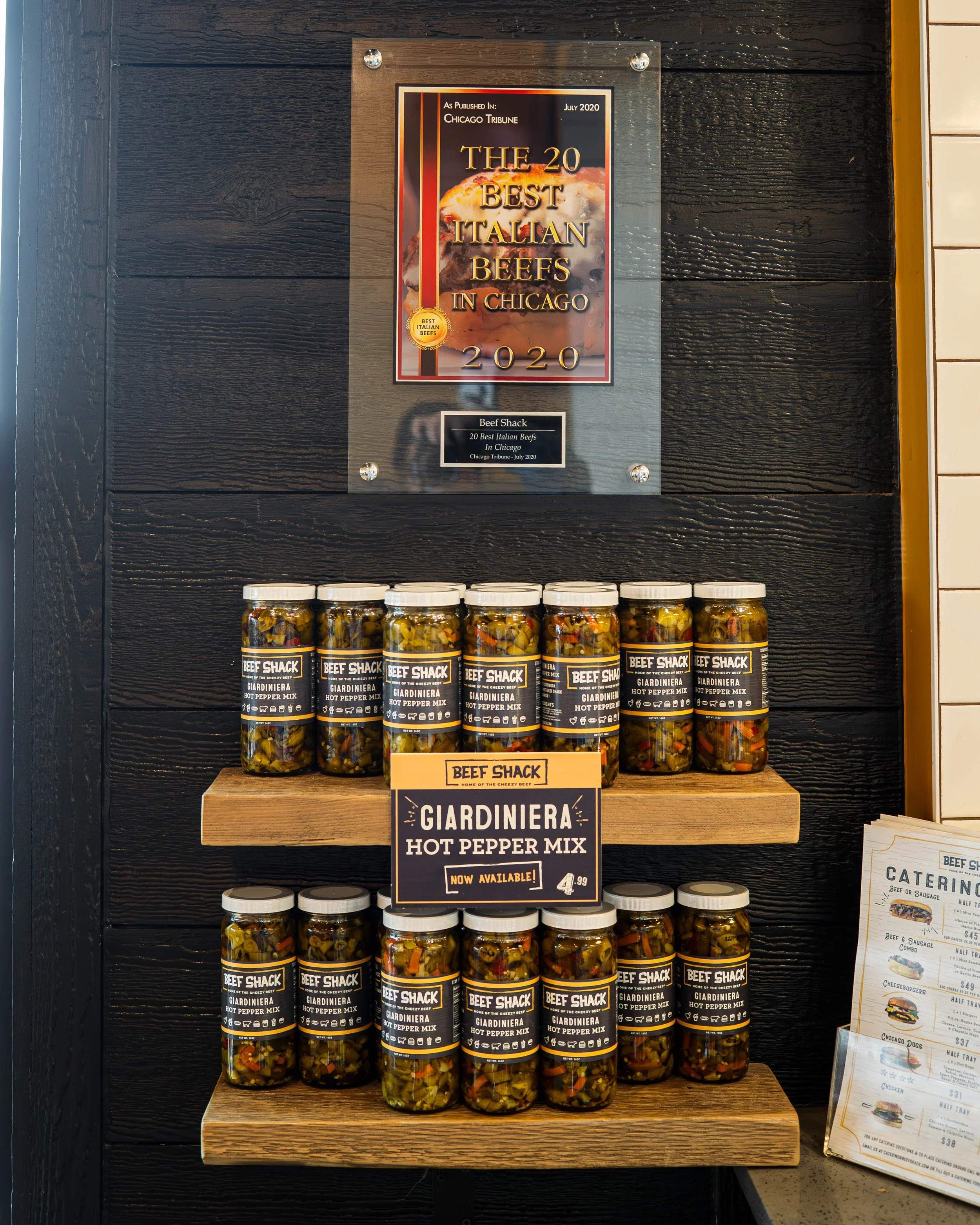 Display of jars of Giardiniera hot pepper mix from Beef Shack on wooden shelves with a poster about the best Italian beefs in Chicago above.