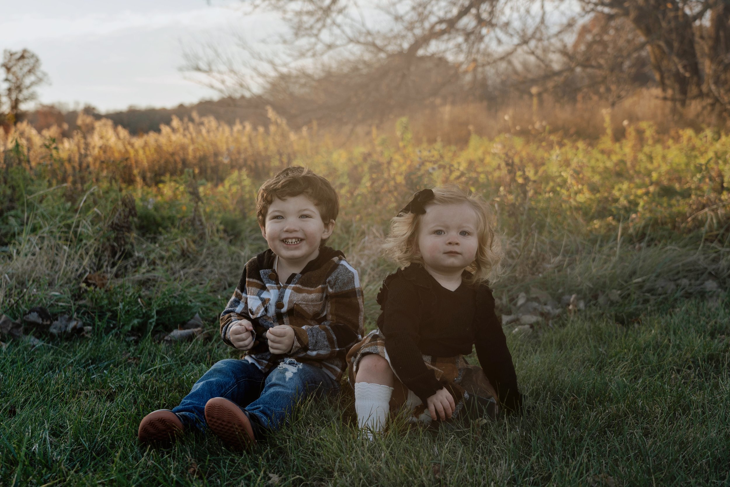 Two young children, a boy and a girl, sitting on grass outdoors during autumn, with trees and golden foliage in the background.