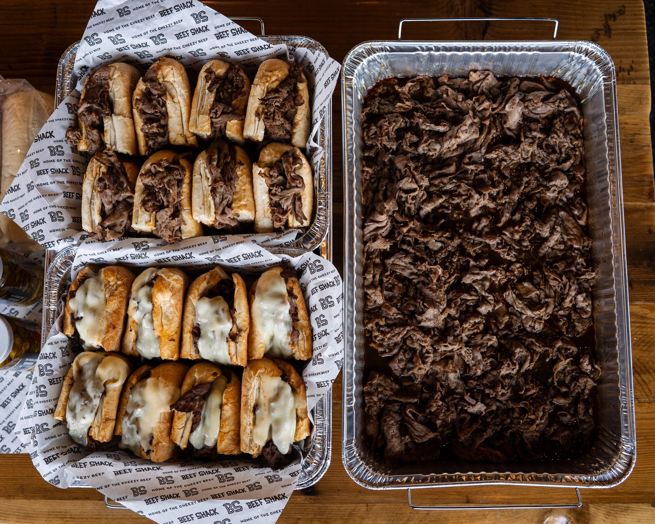 Aluminum trays filled with barbecue beef sandwiches, some topped with cheese, and shredded cooked beef on the side, on a wooden table.