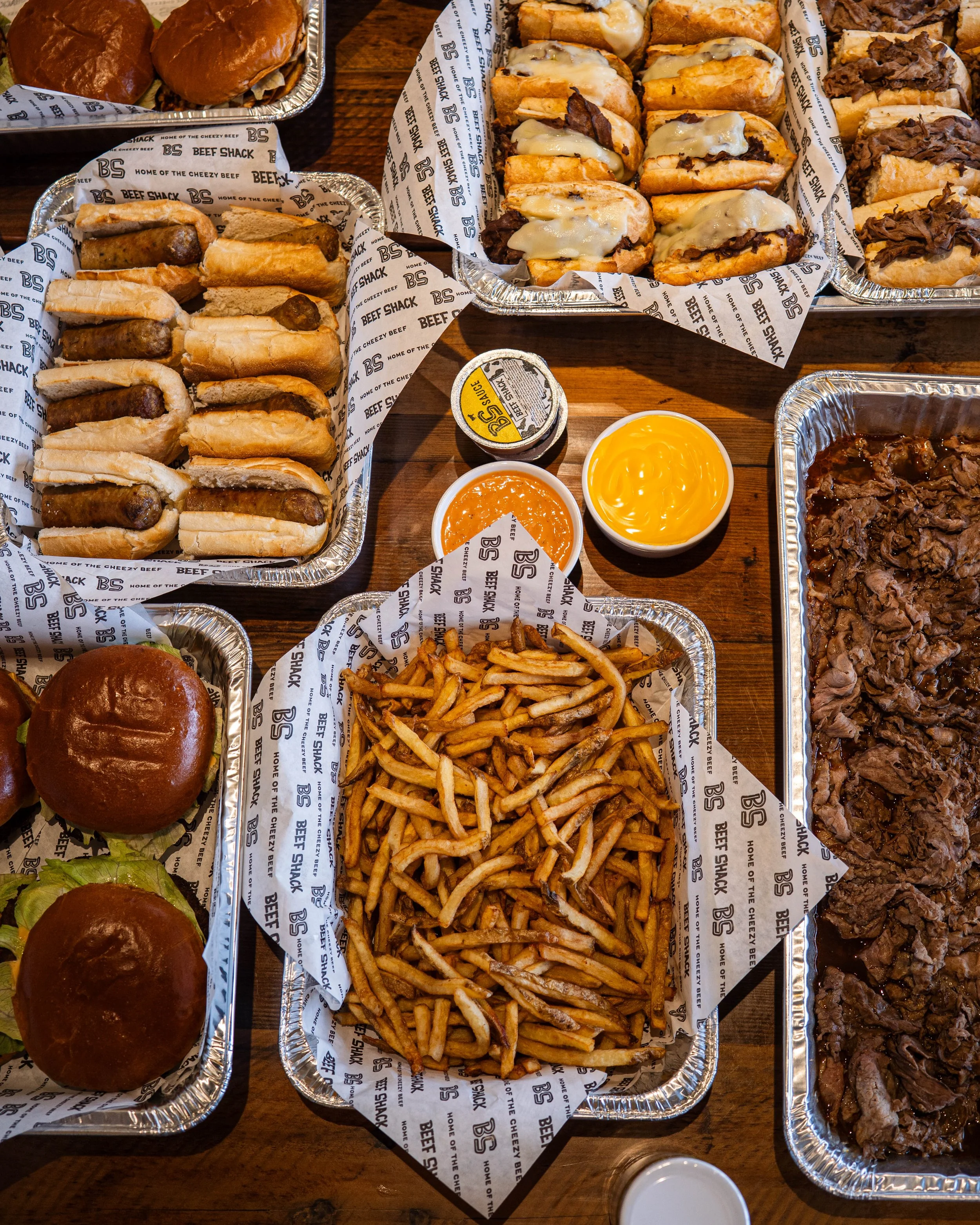 An assortment of fast food including sliders, French fries, small containers of cheese sauce, barbecue, baked beans, and mini burgers on a wooden table.