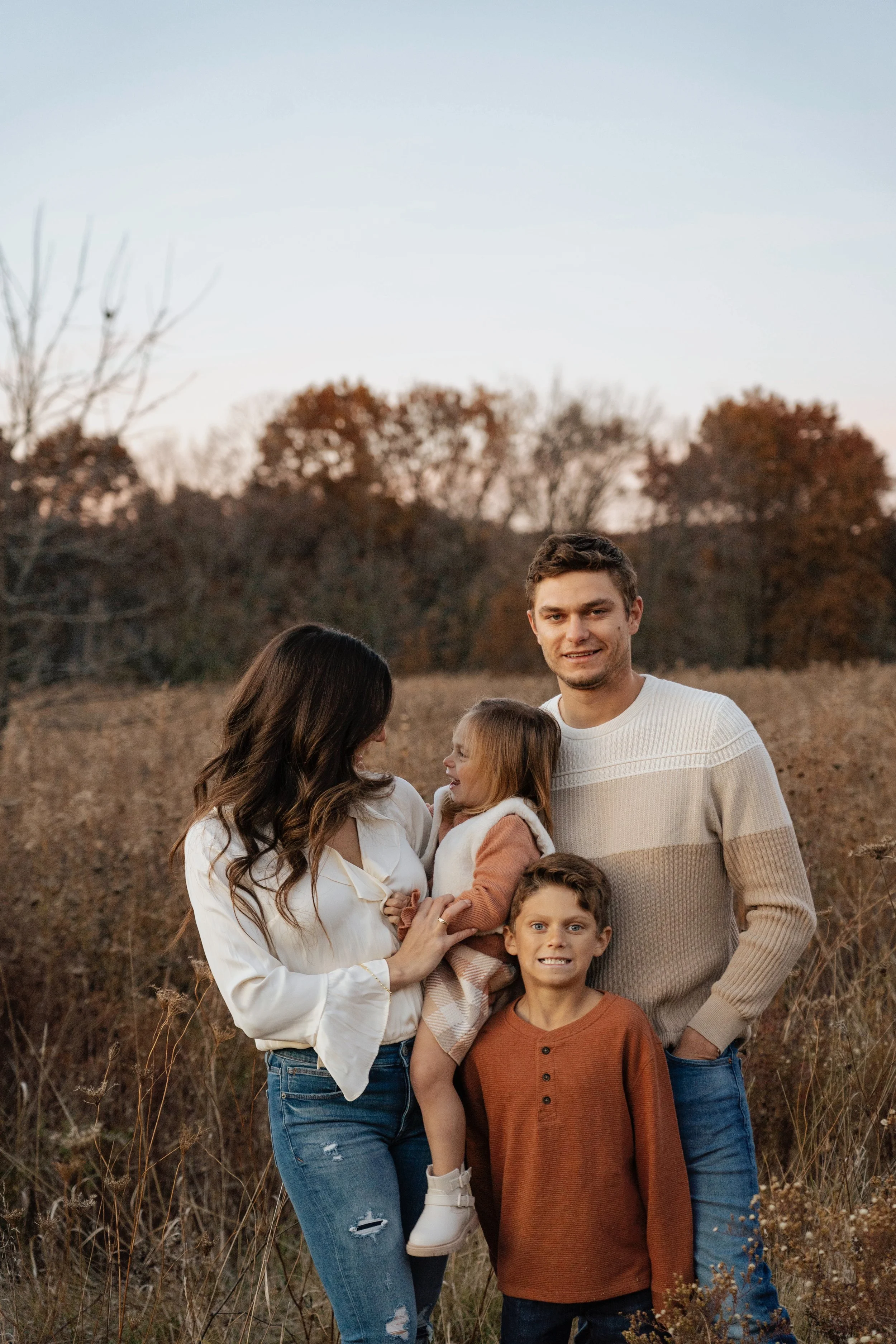 A family of five standing outdoors in an autumn landscape, including a mother, father, two children, and a young girl being held by the mother, all smiling and dressed in fall clothing.