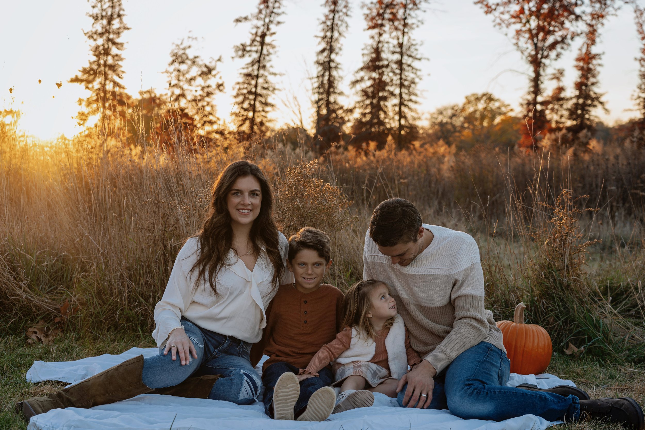 A family of five sitting on a blanket outdoors during sunset with trees in the background. There is a pumpkin nearby.