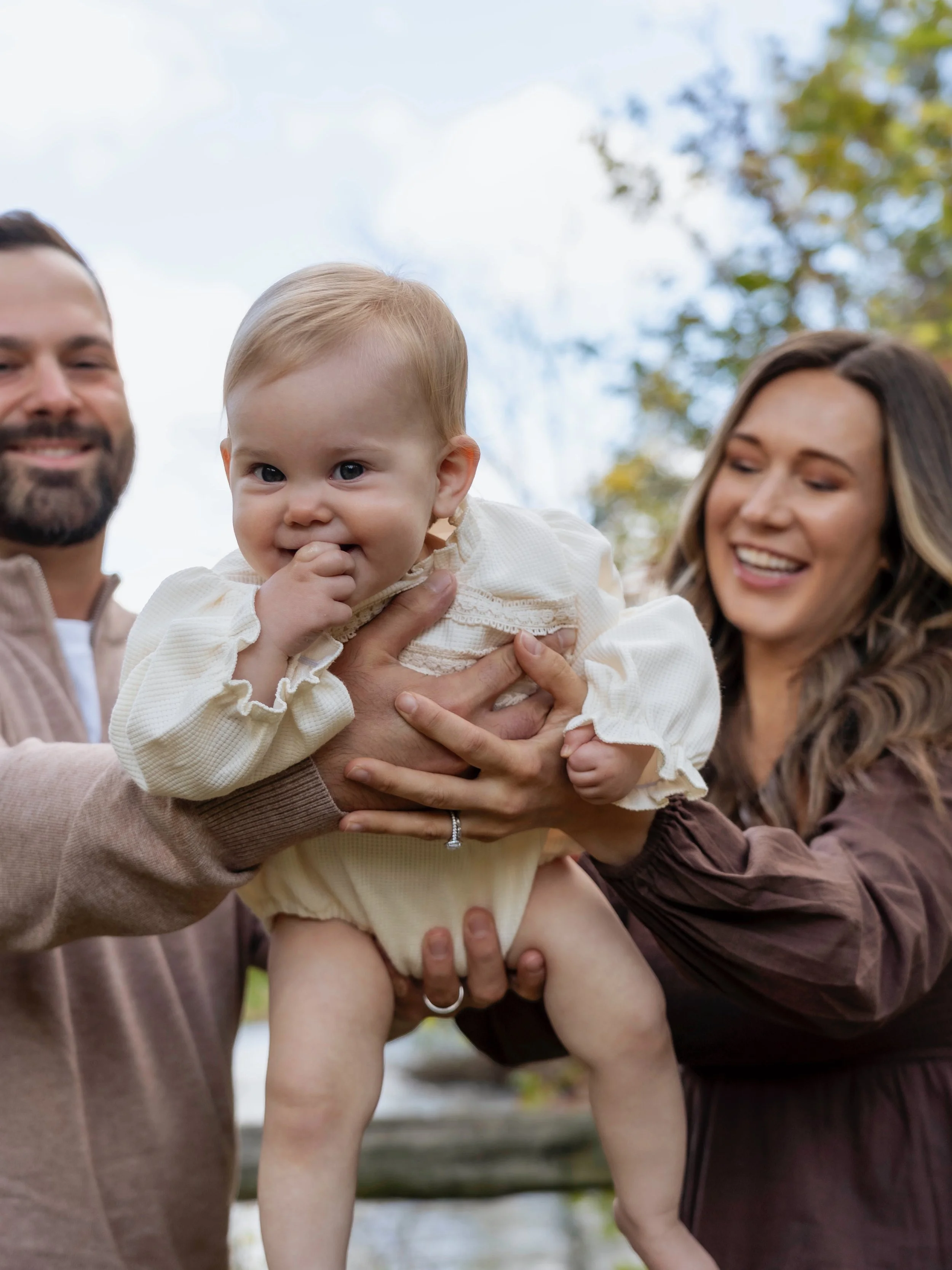 A happy baby being held up by a man and woman outdoors with trees and sky in the background.