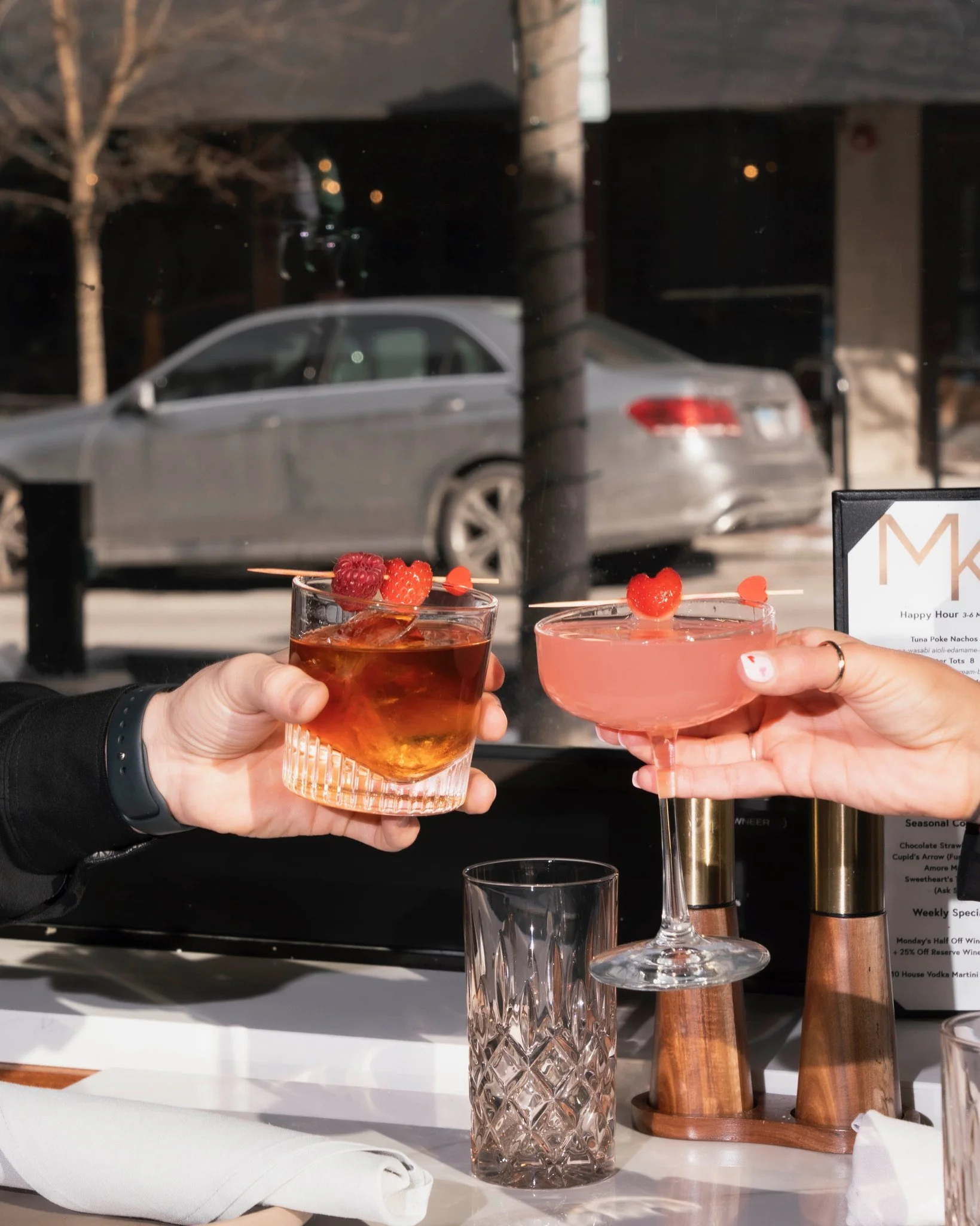 Two people holding cocktails at a restaurant table, with a car outside the window in the background.