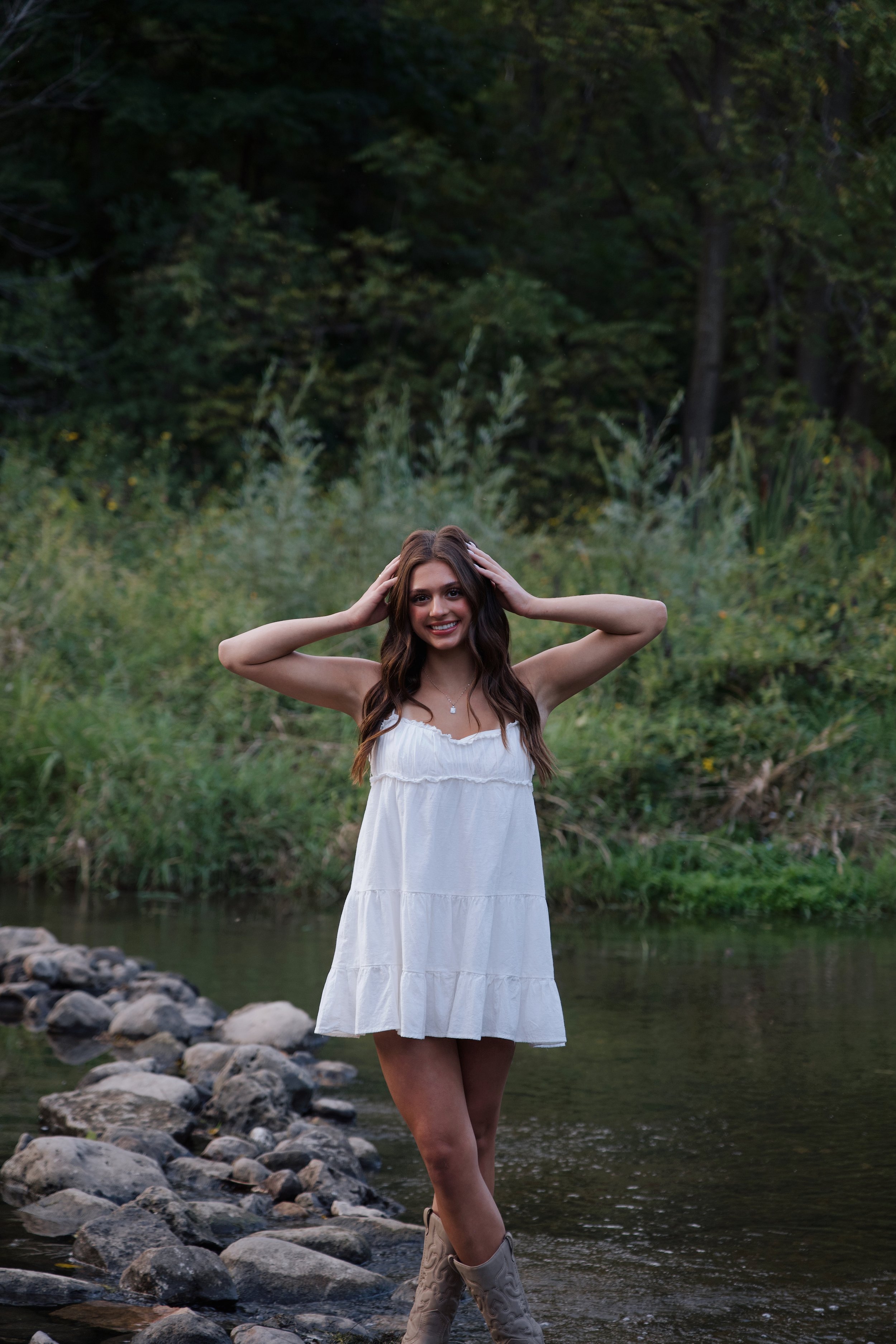 A young woman with long brown hair wearing a white sleeveless dress and cowboy boots standing on rocks in a creek, smiling with her hands on her head, surrounded by greenery.