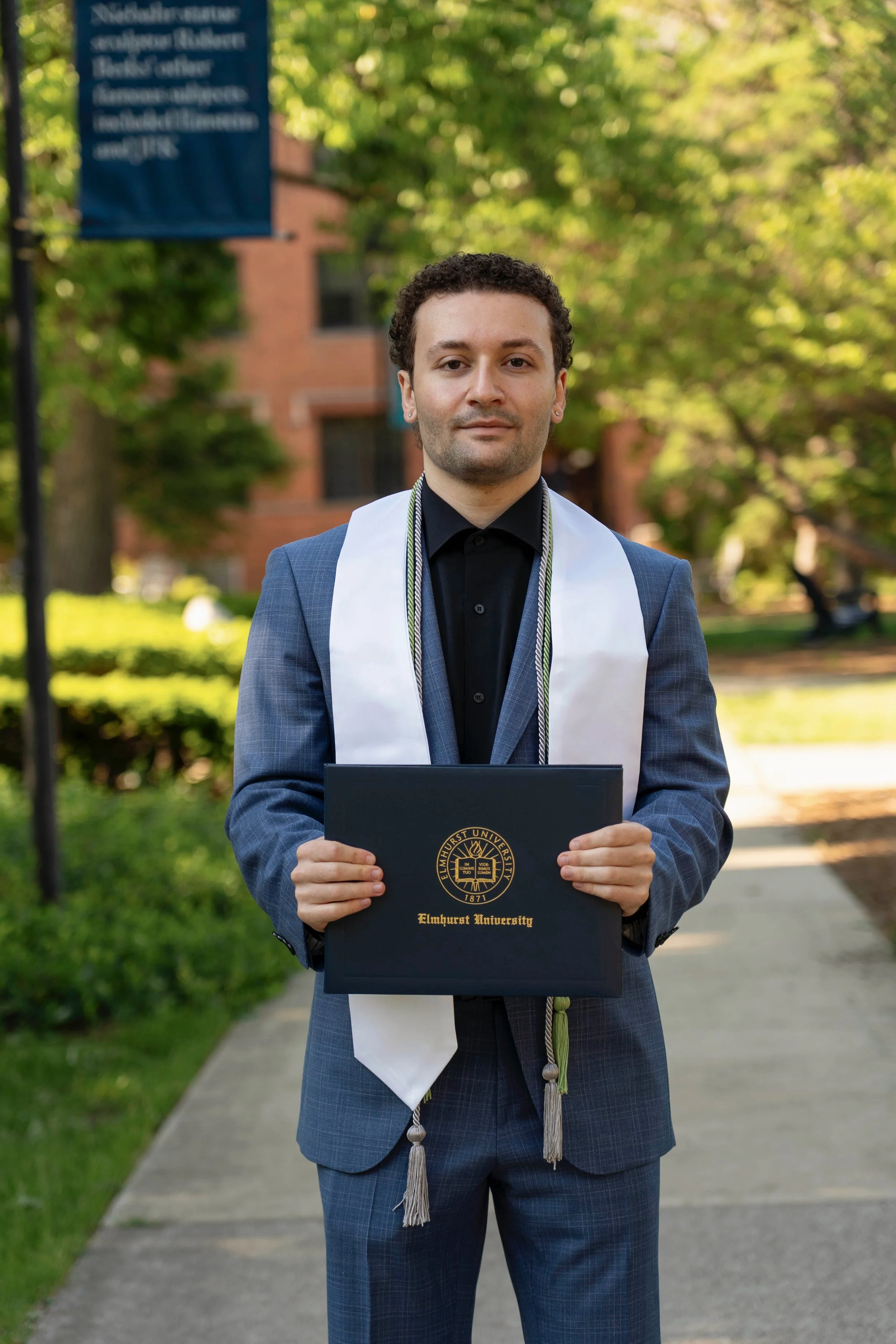 Graduate in a suit holding a diploma from Elmhurst University, standing outdoors on a sidewalk surrounded by trees and greenery.