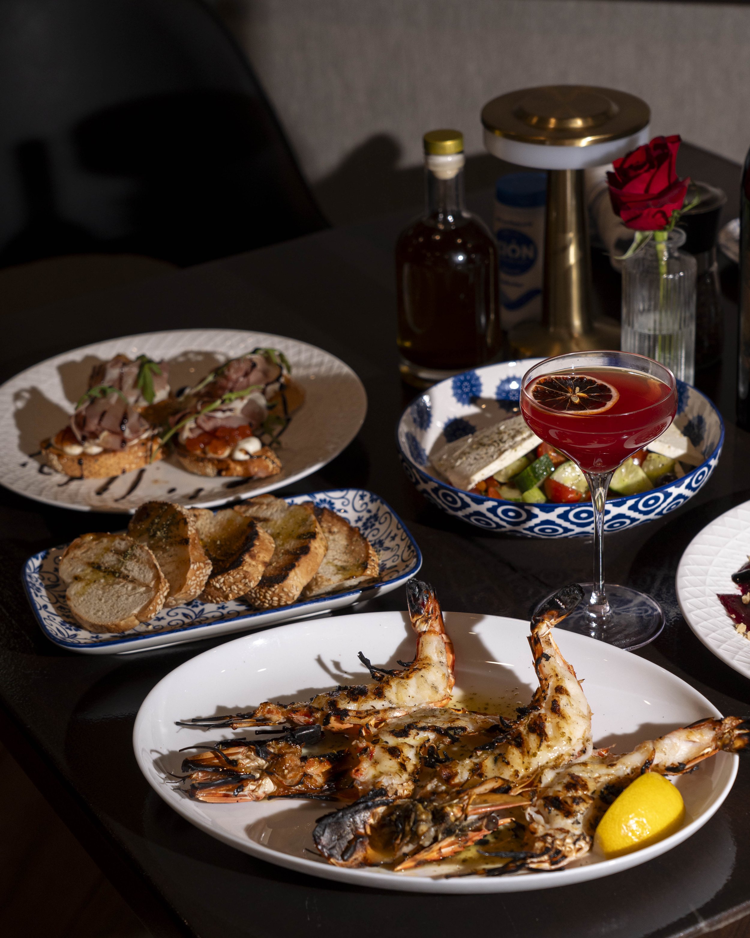 Seafood dinner with grilled prawns, slices of toasted baguette, a salad, a cocktail, and various condiments on a dark table.