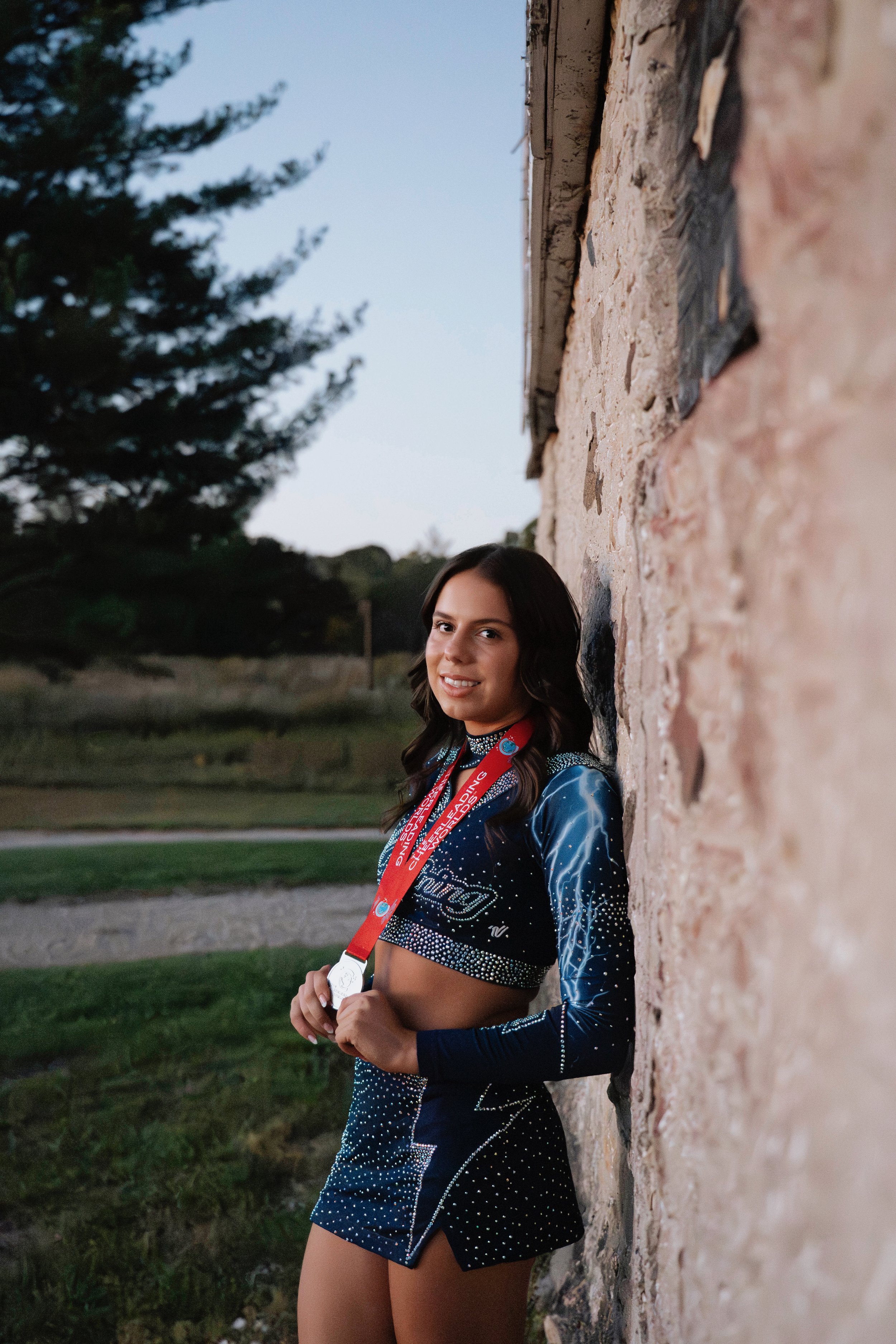 Young female cheerleader with medals, wearing a navy blue sparkly cheerleading uniform, standing against a stone wall outdoors at dusk.