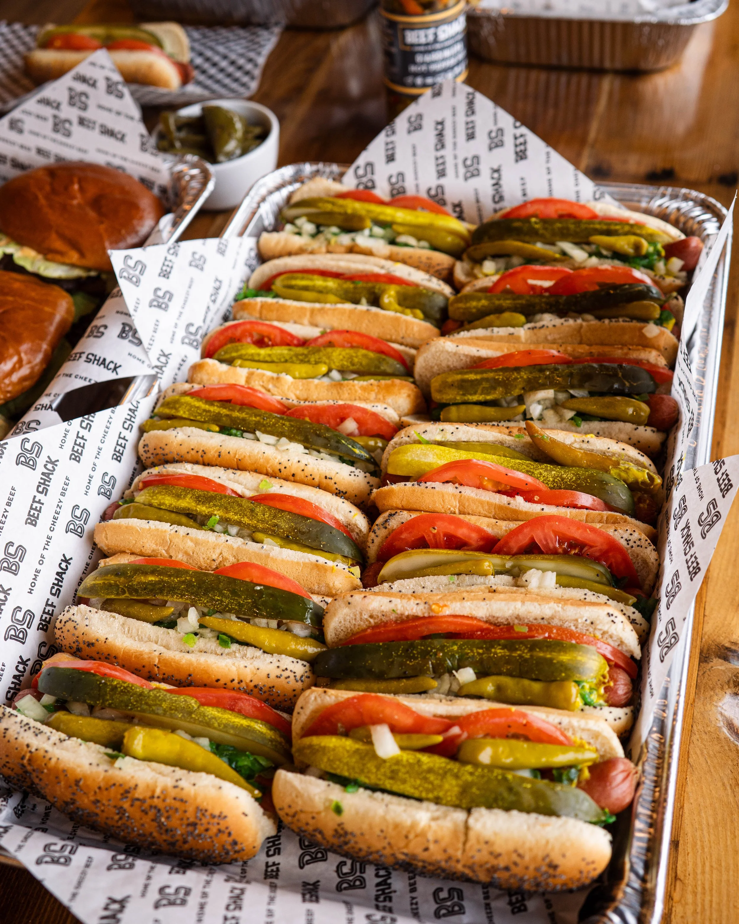 A tray of hot dogs topped with sliced pickles, tomatoes, and chopped onions, lined with branded paper, on a wooden table.