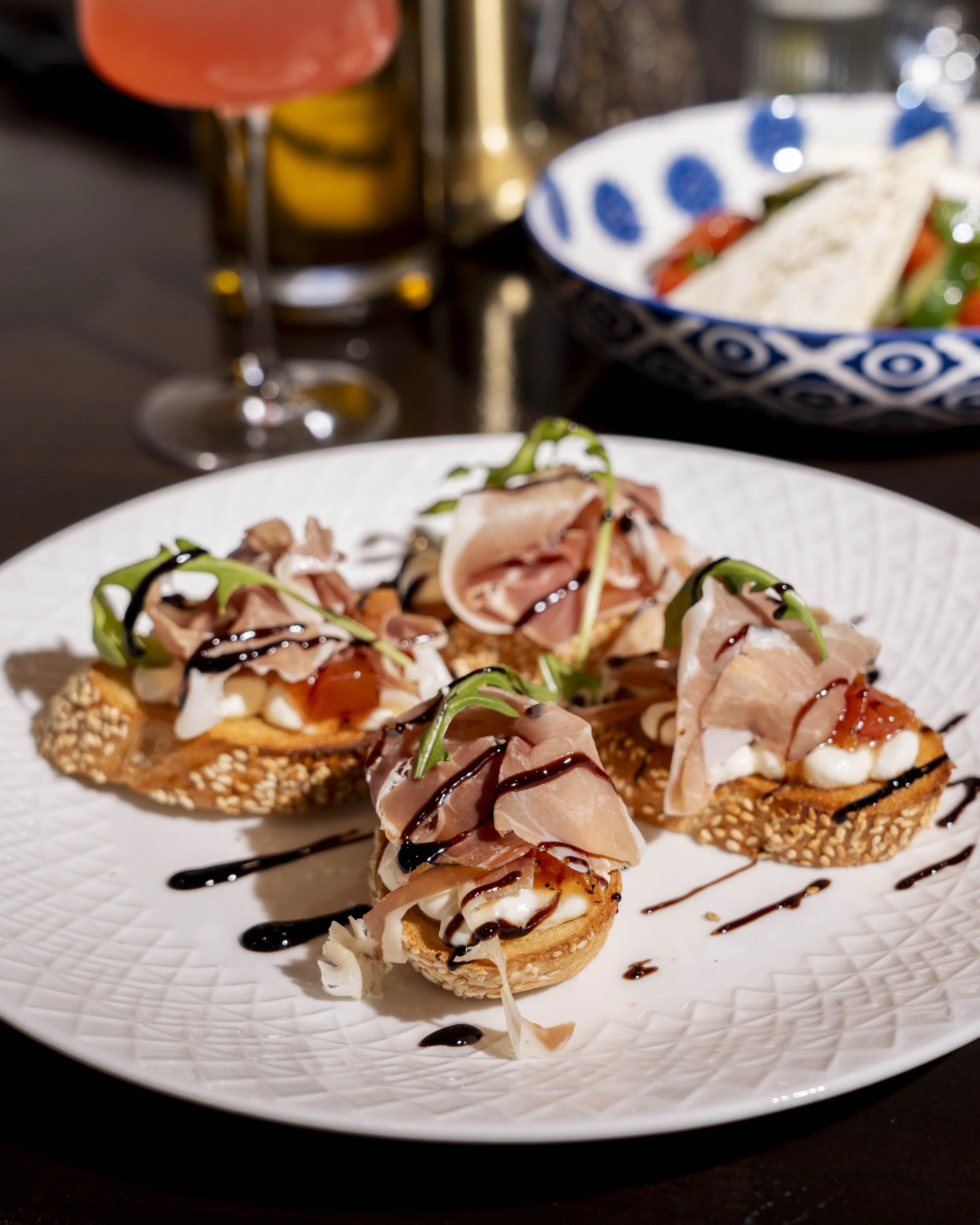 Appetizer of toasted bread slices topped with prosciutto, cheese, greens, and drizzled with balsamic glaze on a white plate.