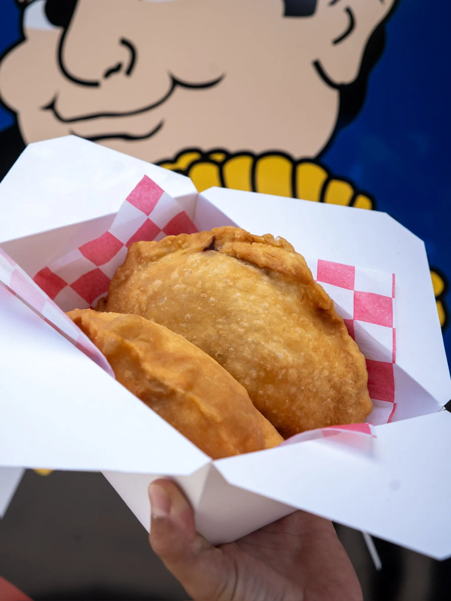 Close-up of a hand holding a white paper tray with two fried chicken pieces, lined with red checkered paper, with a cartoon character with glasses and a yellow shirt in the background.