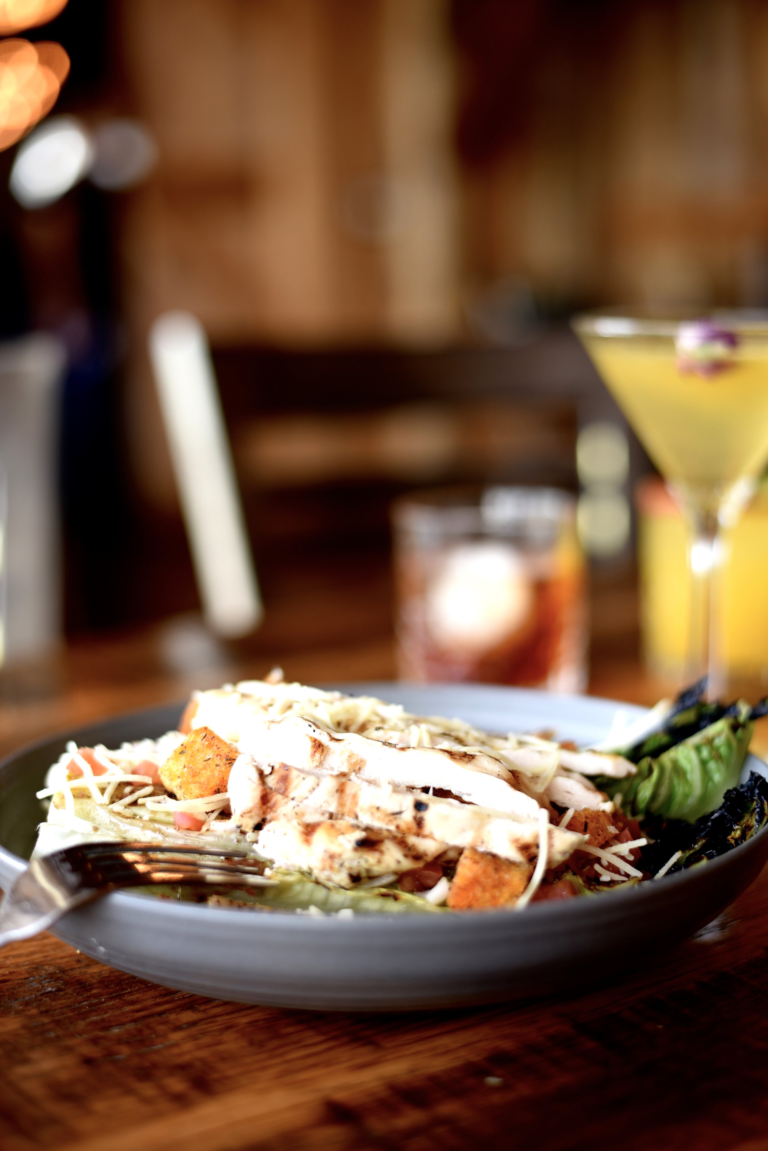 A plate of grilled chicken Caesar salad with shredded cheese, croutons, lettuce, and grilled asparagus. In the background, there is a glass of dark soda, a cocktail, and a blurred bar or restaurant setting.