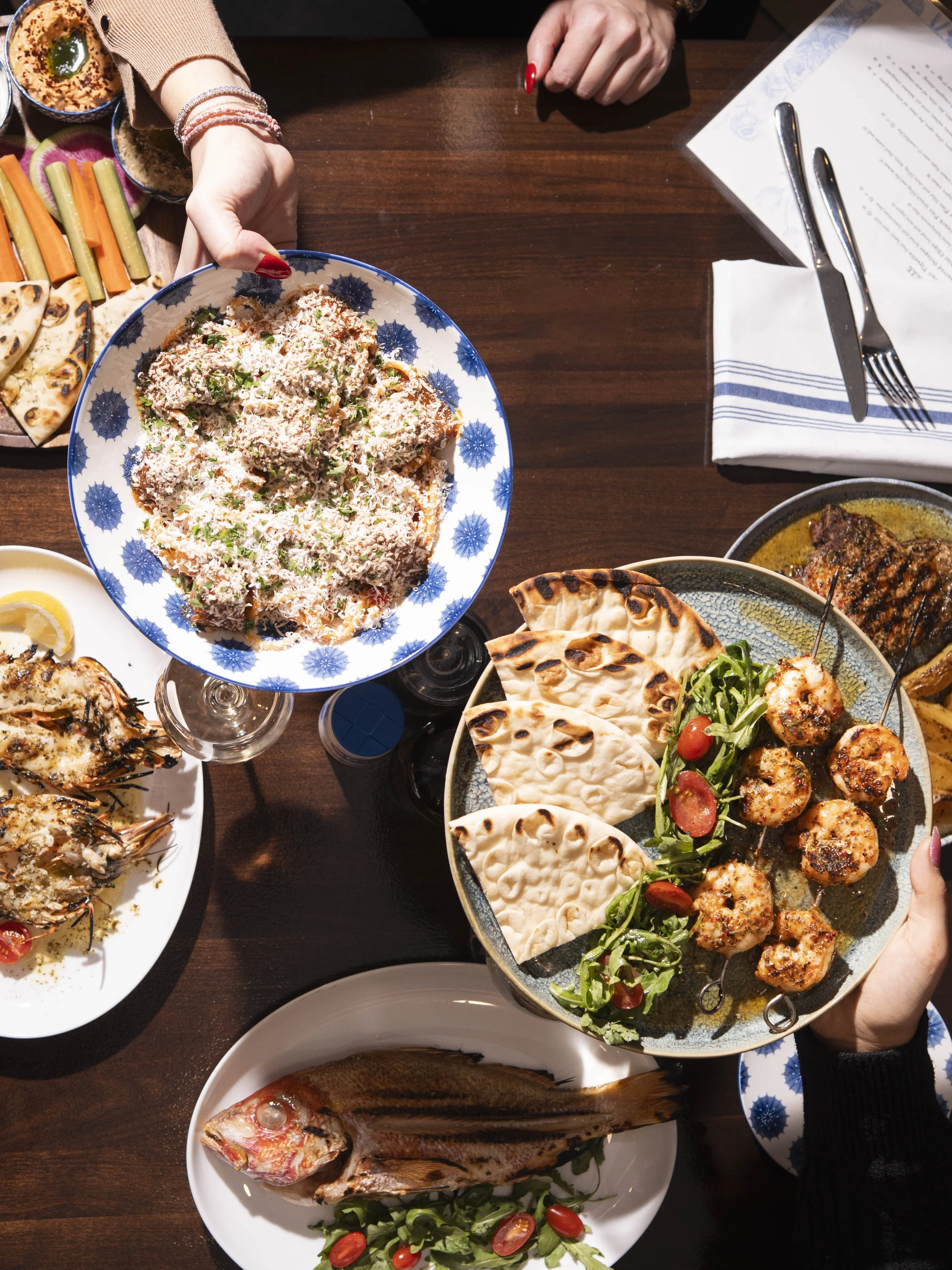 A high-angle view of a table with various dishes, including a plate of rice topped with shredded coconut, a bowl of grilled chicken skewers with pita bread and greens, a whole cooked fish garnished with herbs, and a platter of grilled vegetables and 