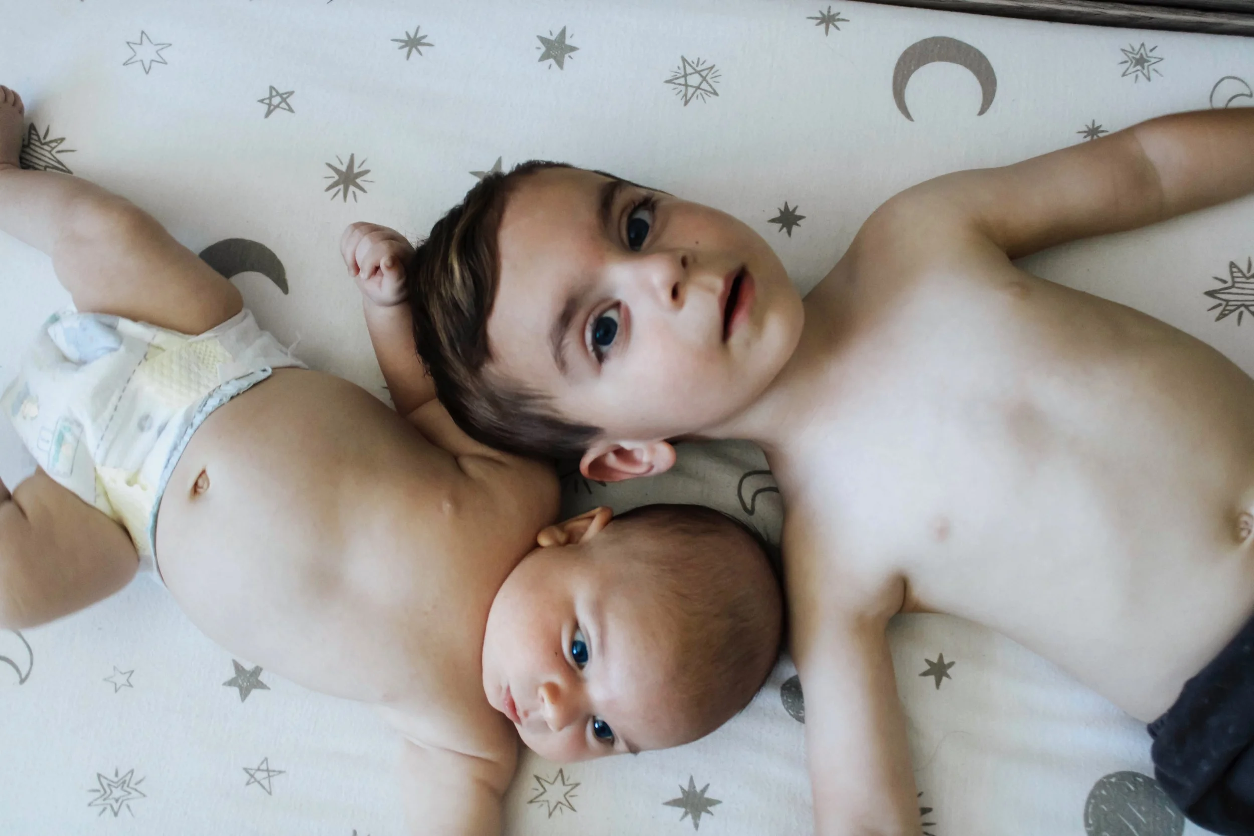 Two young boys lying on a bed with a star and moon pattern, both shirtless, looking up at the camera.