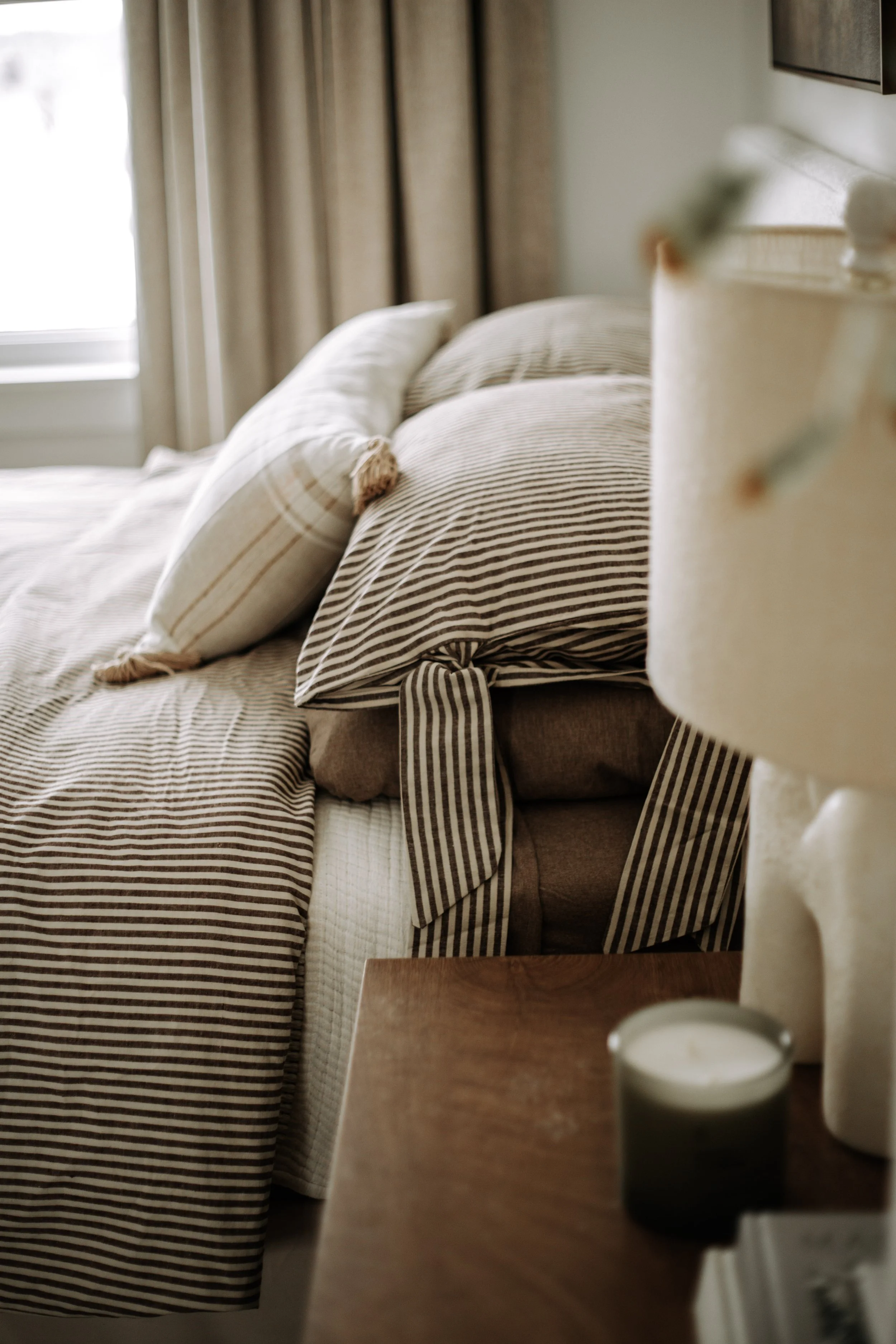 Wood bedroom nightstand styled with a sculptural lamp and greenery beside layered neutral bedding.