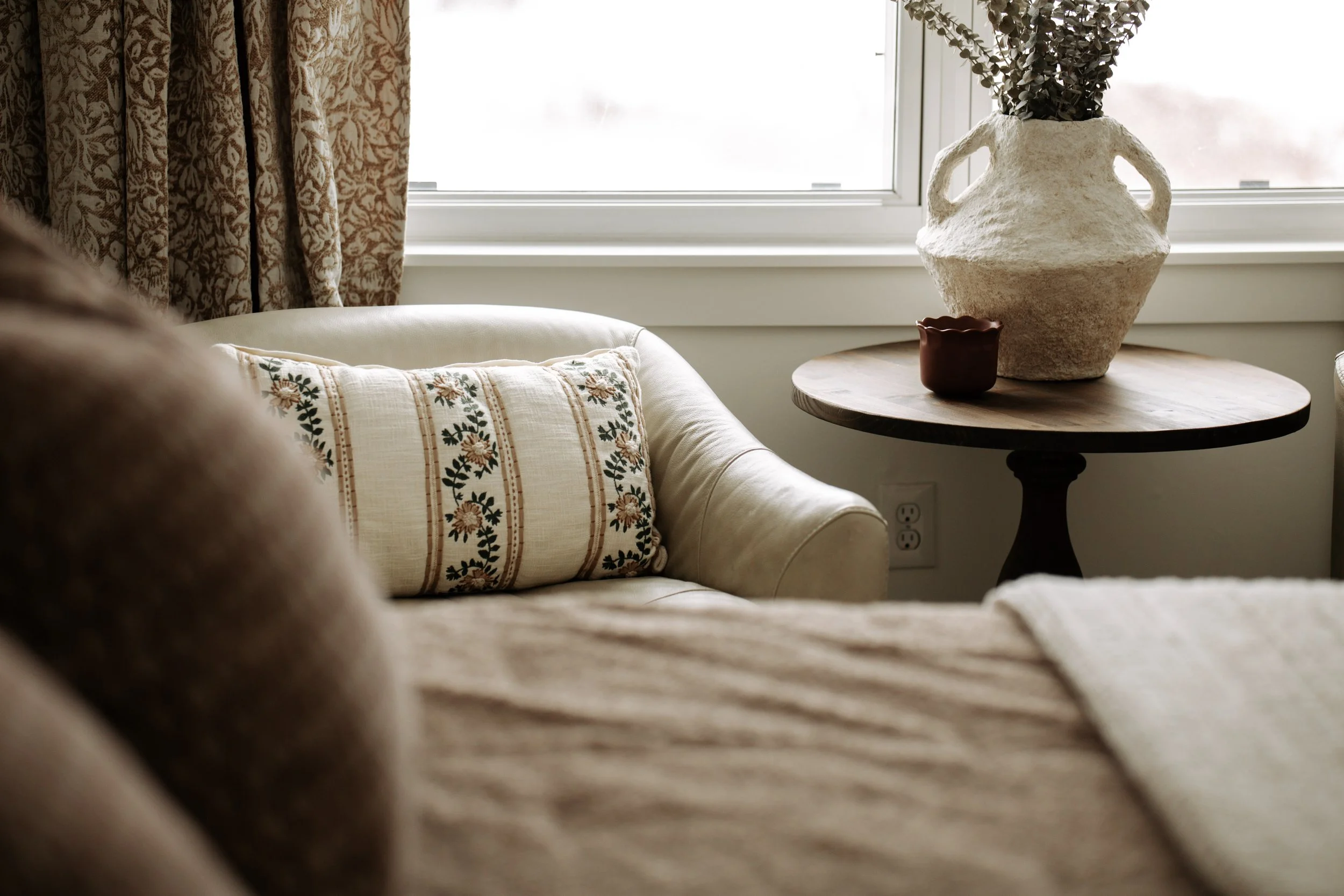 Cozy bedroom reading chair with embroidered pillow, soft throw and patterned window treatments.