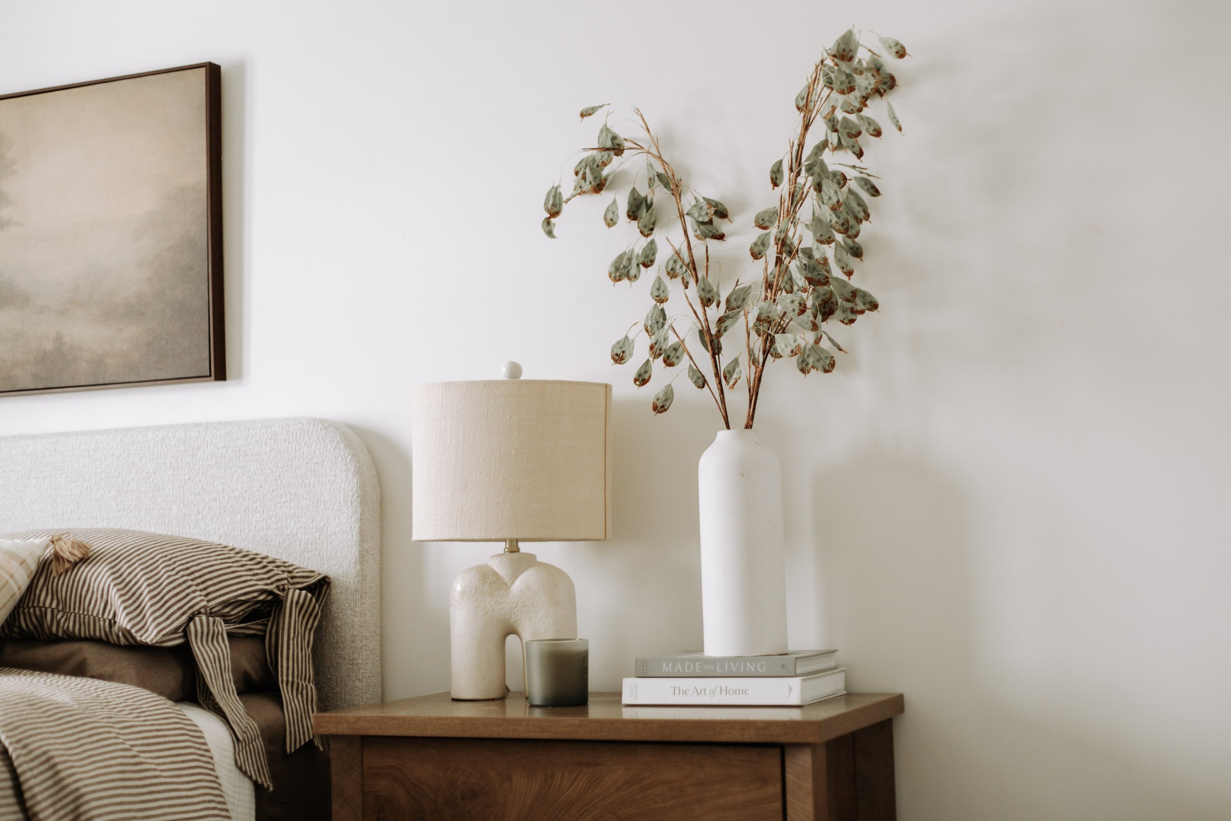 Upholstered headboard by a wood nightstand with  a ceramic vase, architectural lamp and natural greenery.