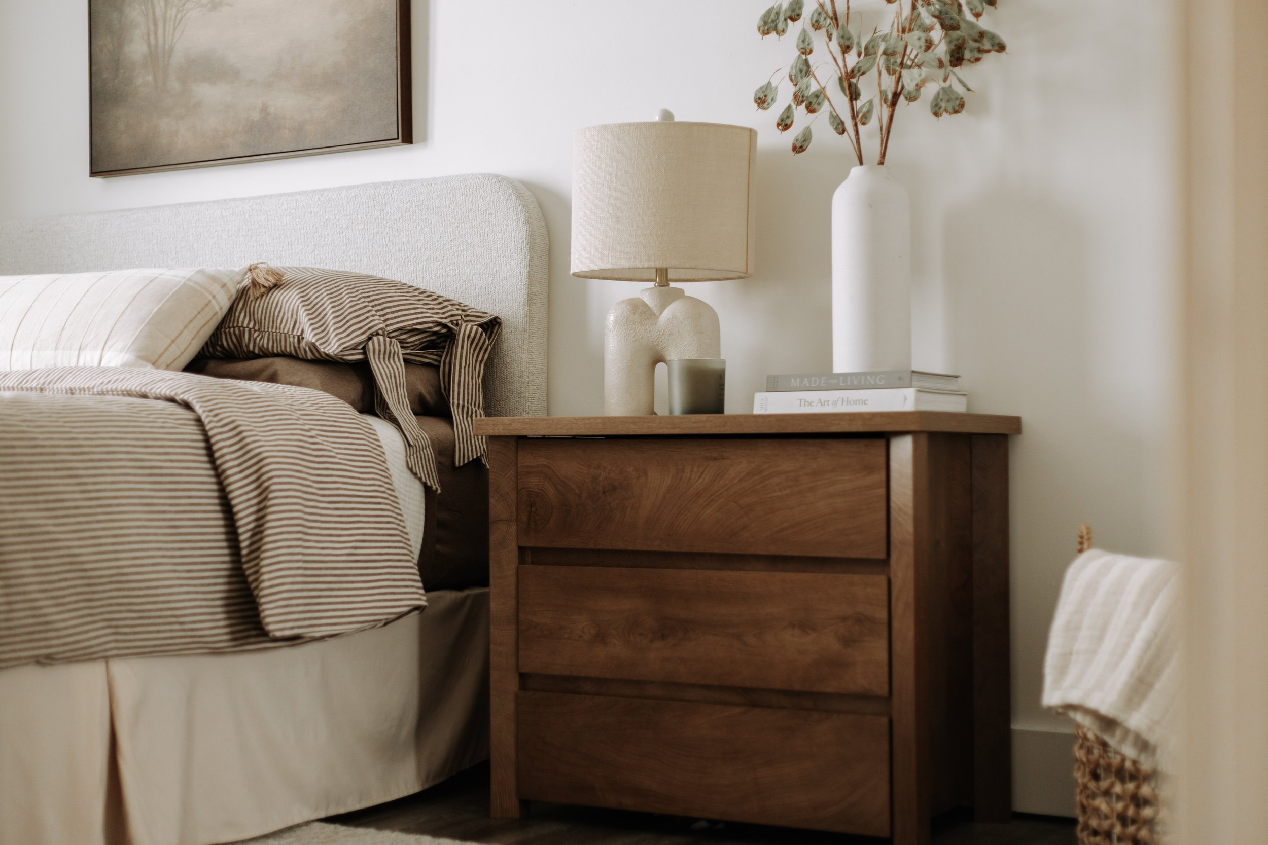 Wood bedroom nightstand styled with a sculptural lamp and greenery beside layered neutral bedding.