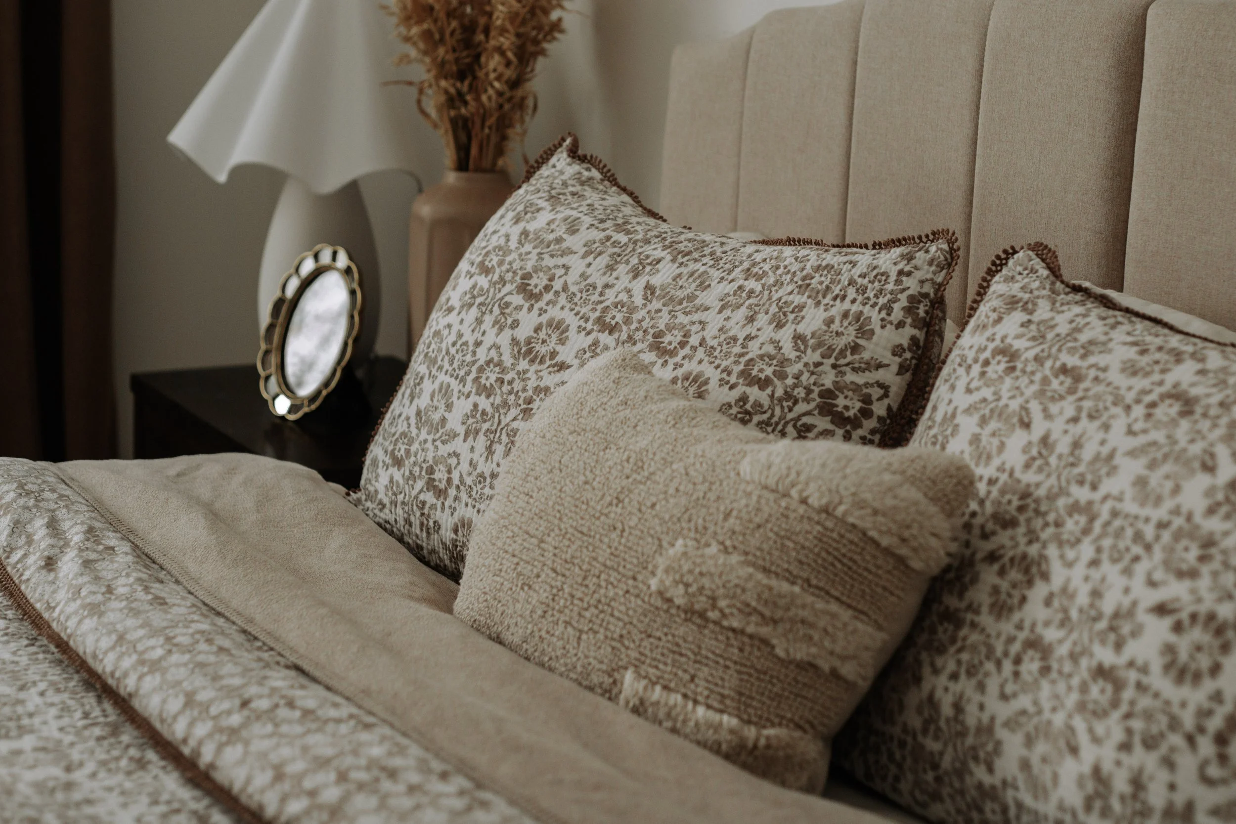 Neutral upholstered headboard with floral pillows, dark wood nightstand and vintage-inspired decor.