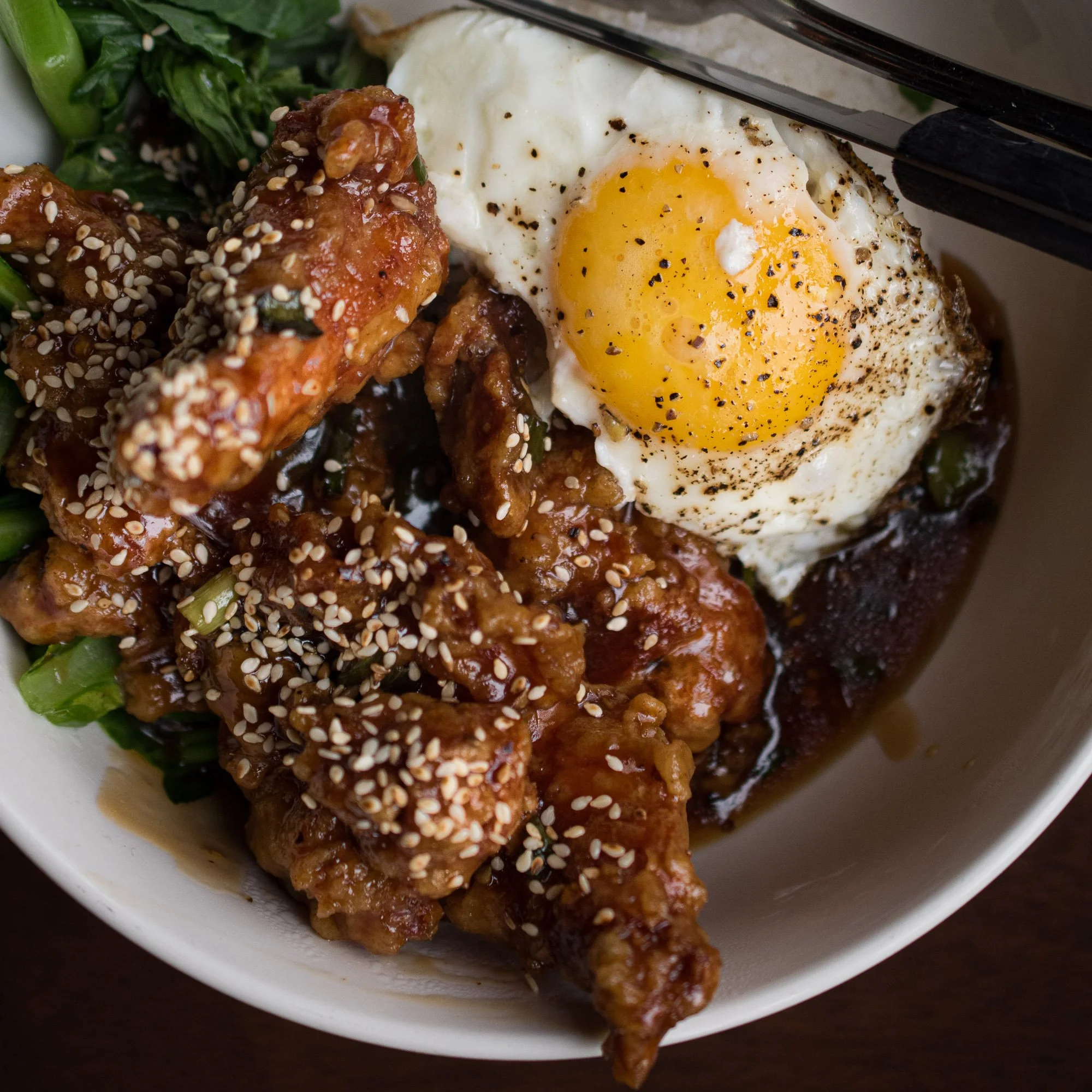 Bowl of Asian-style chicken, green vegetables, and rice topped with sesame seeds, served with a fried egg seasoned with black pepper.