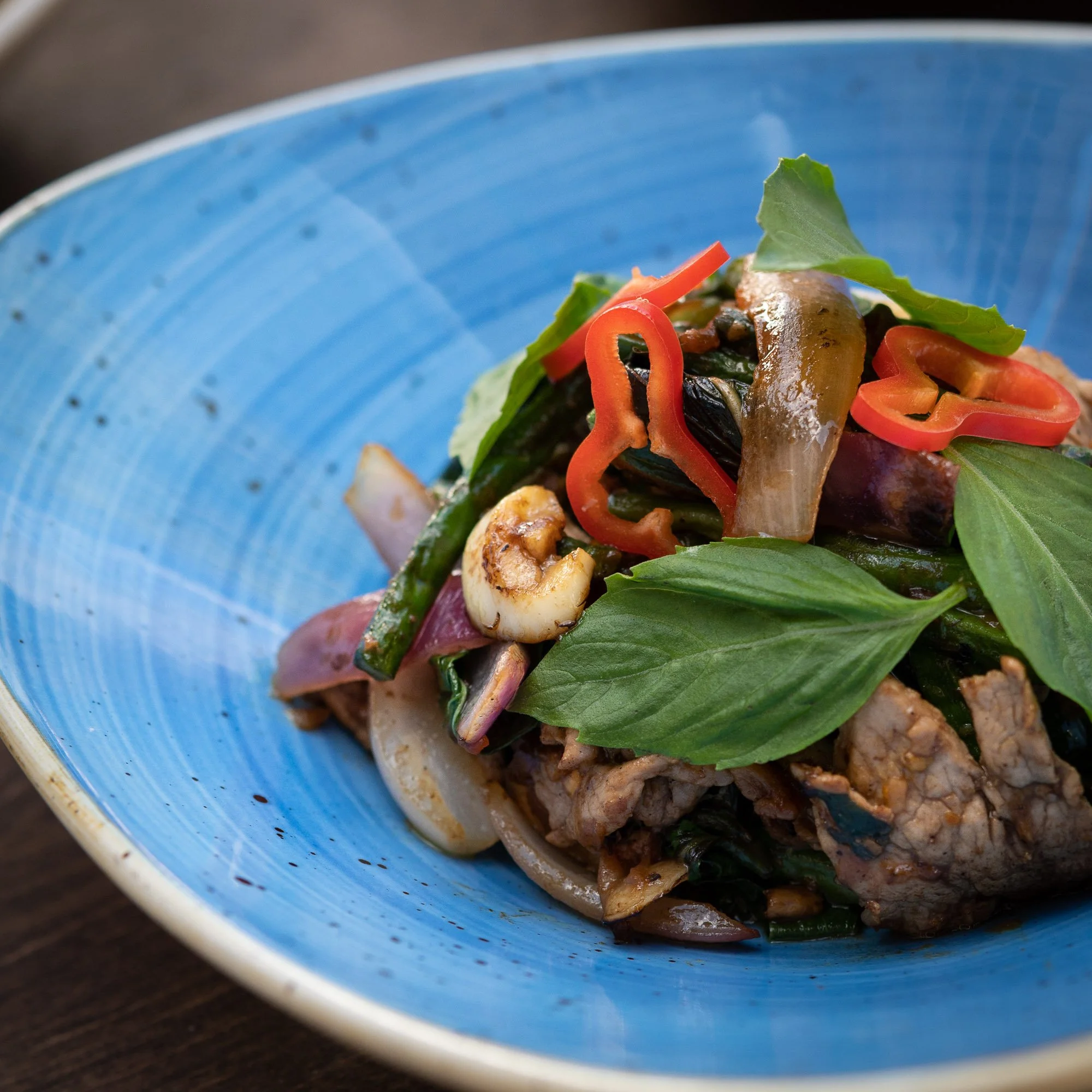 A close-up of a bowl of stir-fried vegetables with basil, red peppers, onions, and pieces of meat on a blue ceramic dish.