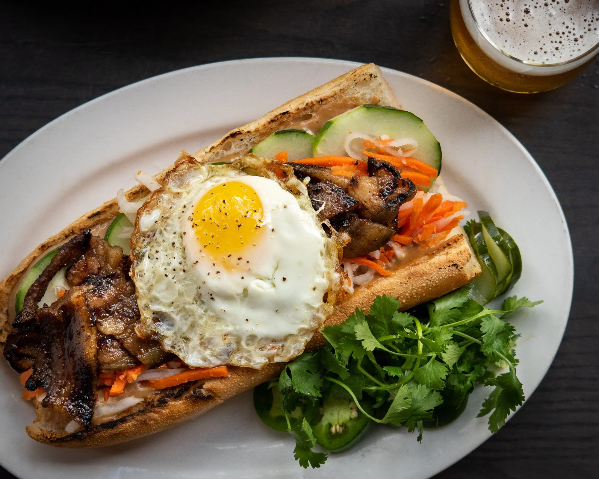 Bánh mì sandwich topped with a fried egg, grilled pork, cucumber slices, shredded carrots, onions, and fresh cilantro, served on a white plate with a side of fresh herbs and a glass of beer in the background.