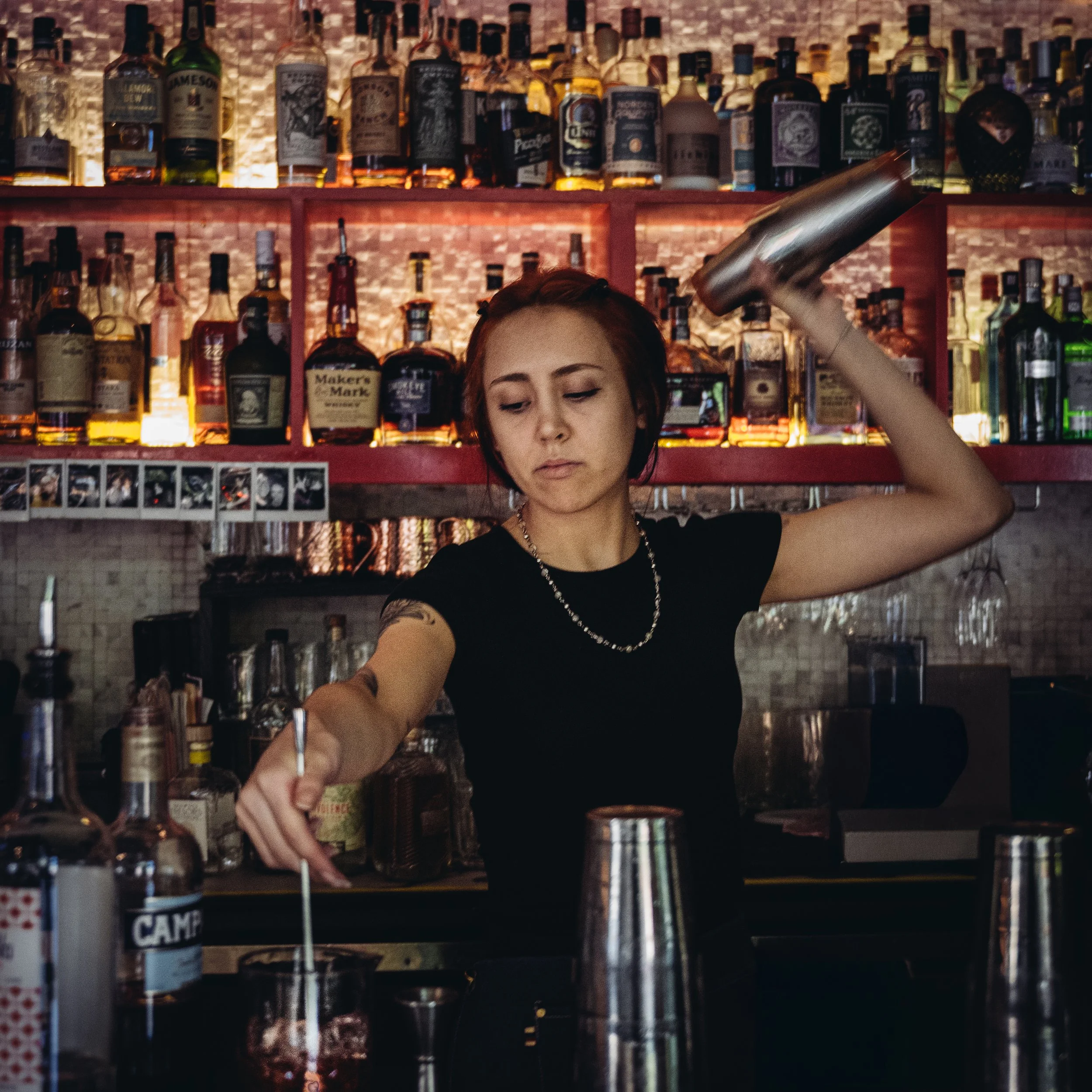 A female bartender with short red hair, wearing a black shirt, pouring a drink with a cocktail shaker behind a bar with bottles of liquor on shelves.