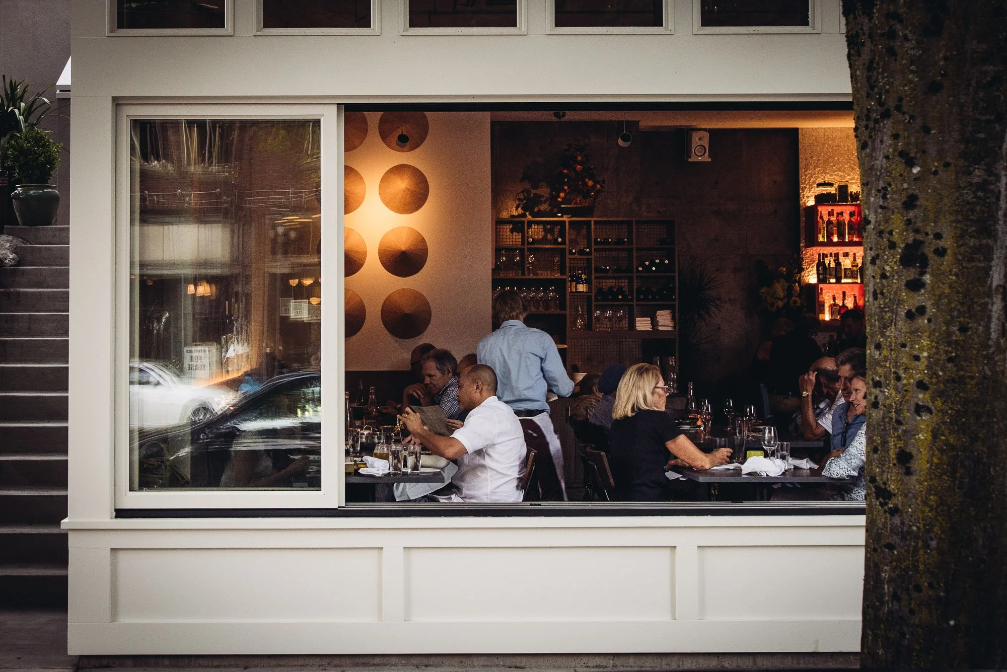 Inside a cozy restaurant viewed through a large window, with diners seated and a waiter serving, illuminated with warm lighting and decorated with wine bottles on shelves.