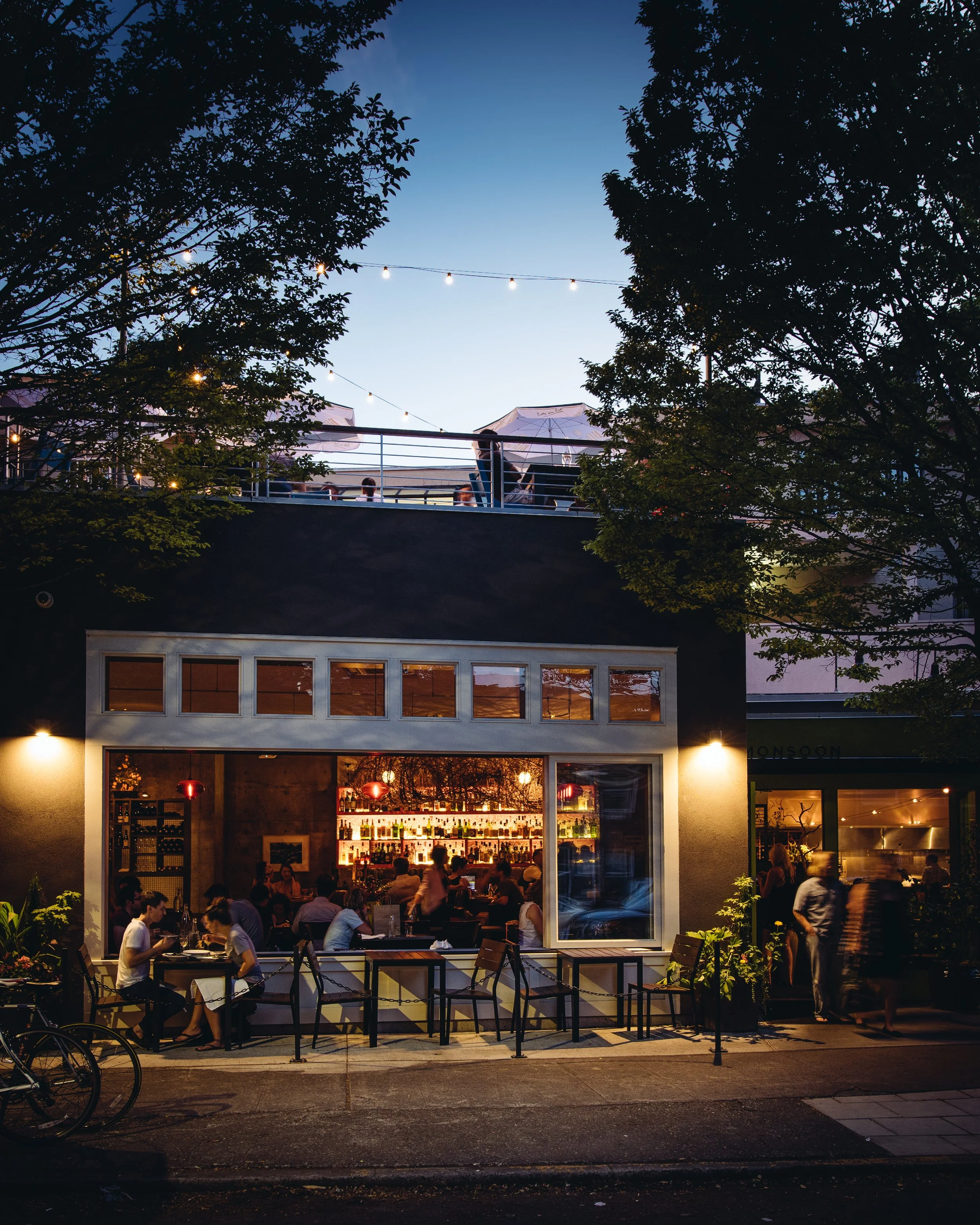 A lively restaurant scene at dusk with people dining inside and outside, illuminated by warm lighting, with a secondary outdoor patio on the roof with umbrellas and string lights, surrounded by trees.