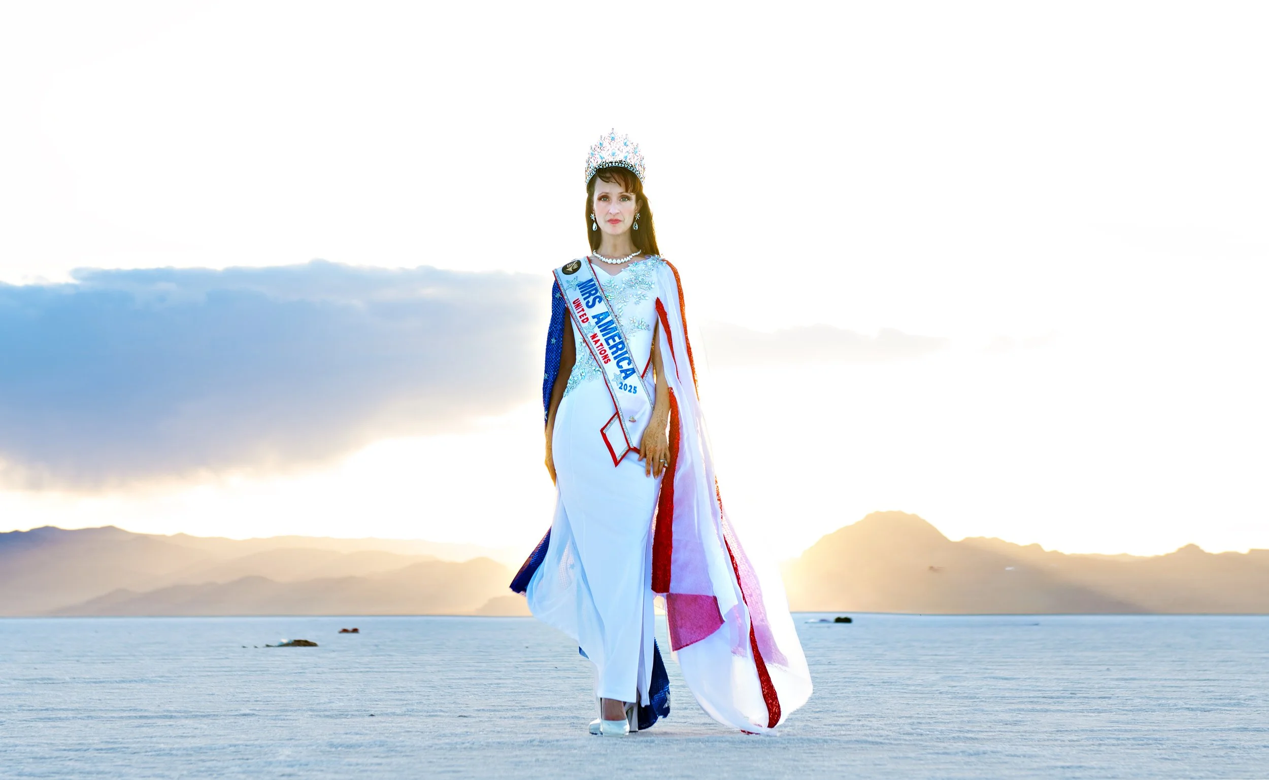 Woman wearing a crown and sash that says "Mrs. America 2025" standing on a salt flat with mountains and a cloudy sky in the background.