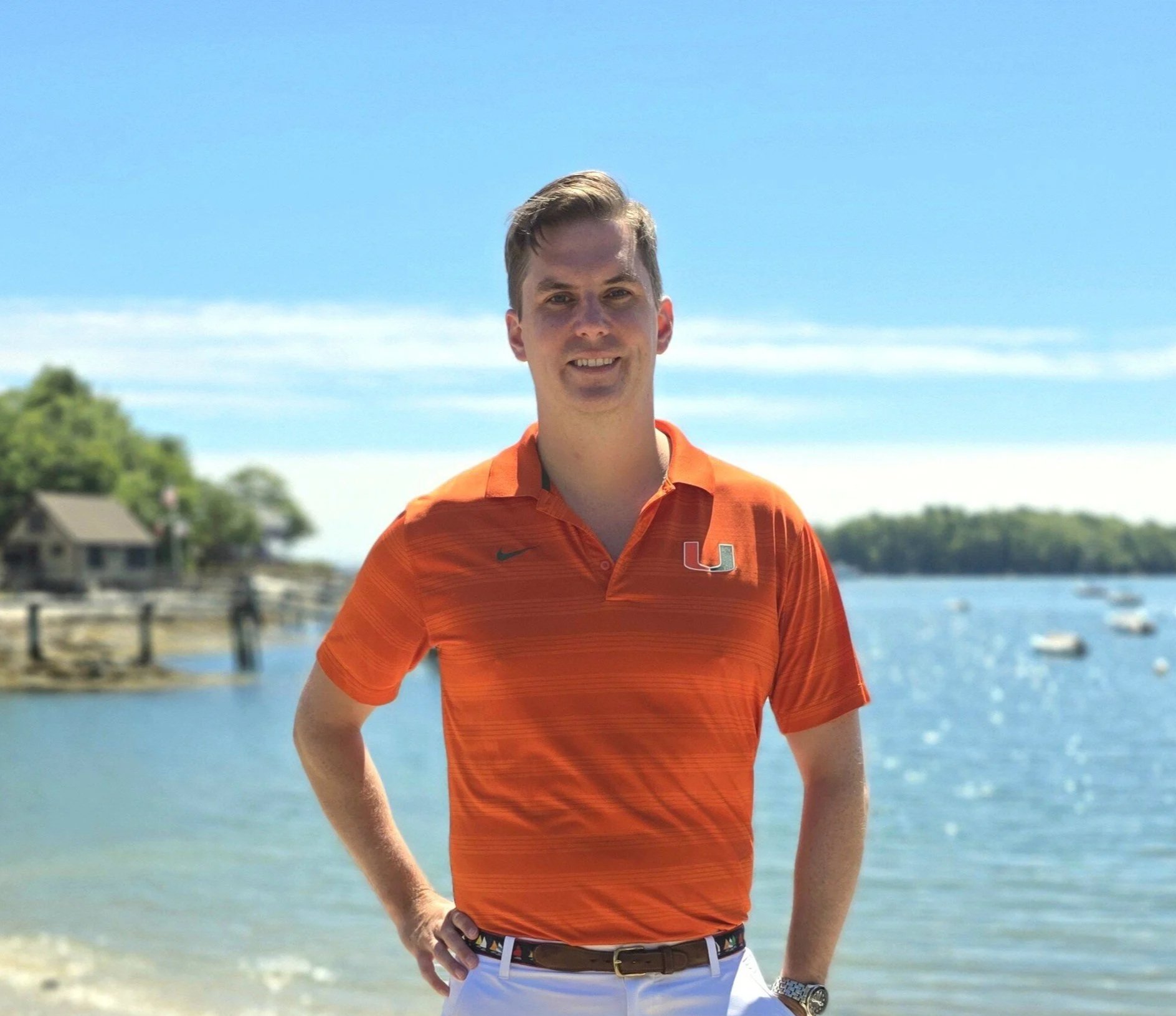A young man standing outdoors near a body of water on a sunny day, wearing an orange Miami Hurricanes polo shirt, with trees and boats in the background.