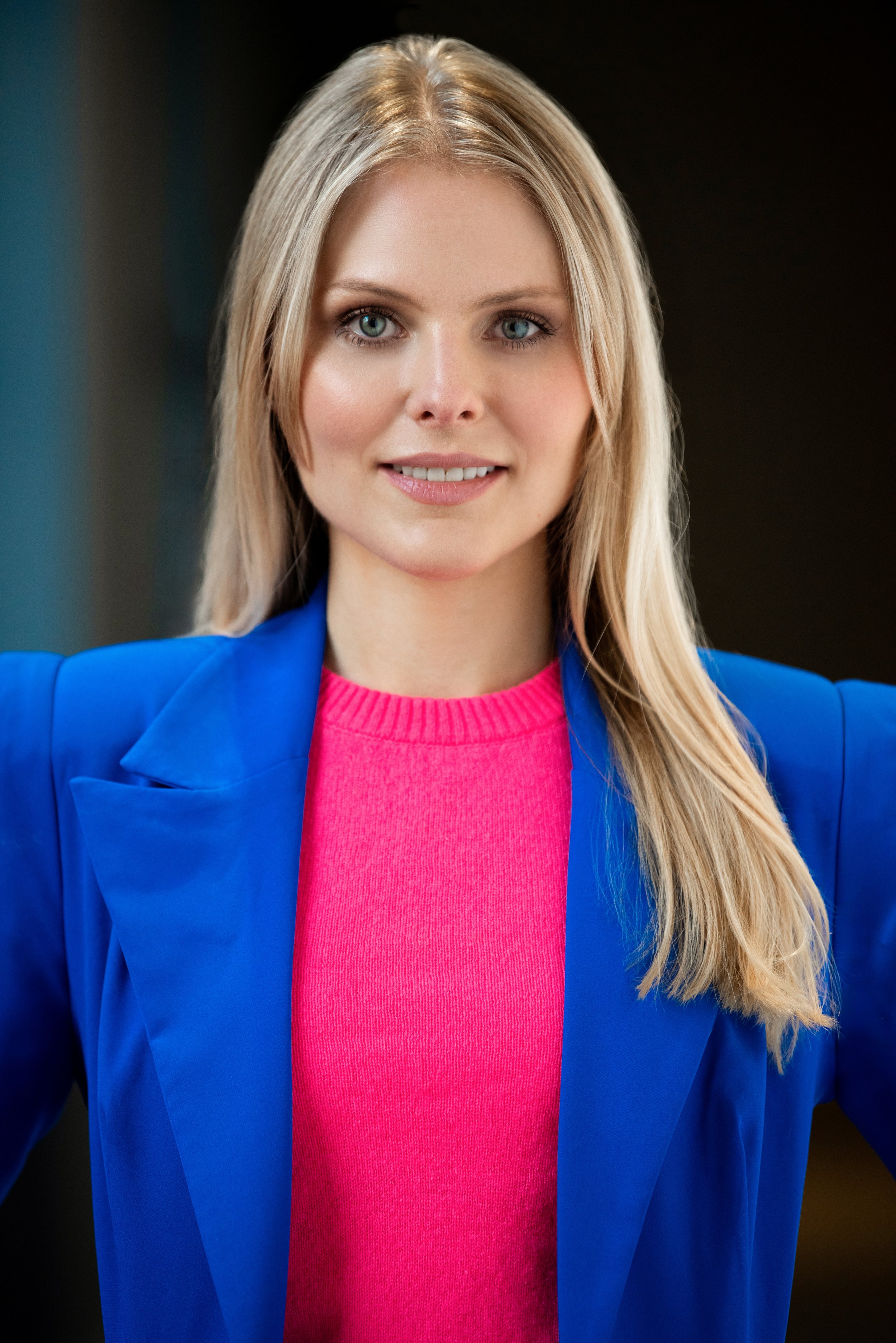 Portrait of a young woman with long blonde hair, blue eyes, wearing a bright pink sweater and a blue blazer, smiling confidently.