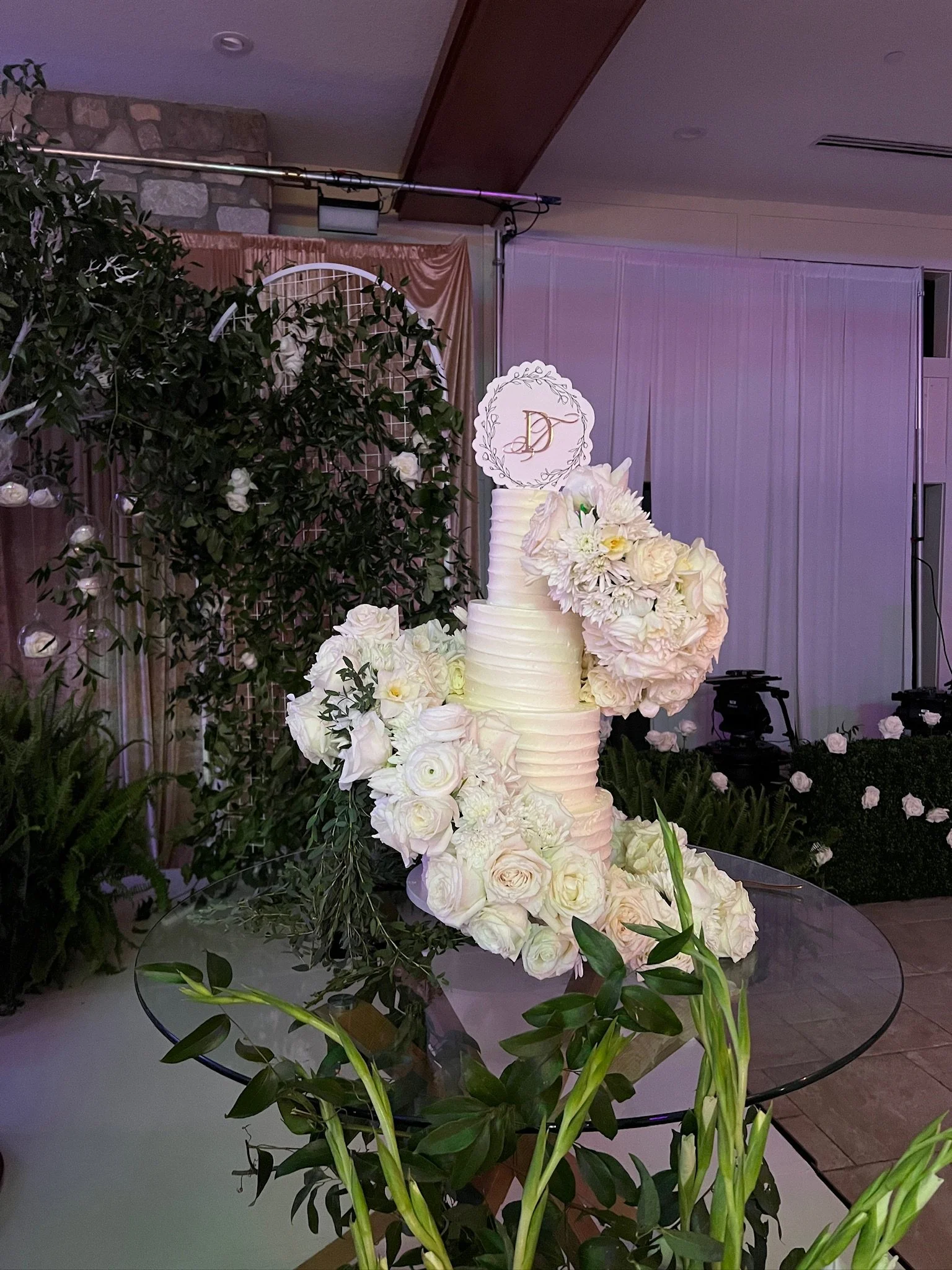 A tall, white, multi-tiered wedding cake decorated with large white flowers, surrounded by green foliage, on a glass table at a wedding or celebration event. From Netflix Love is Blind Daniel and Taylor Wedding.