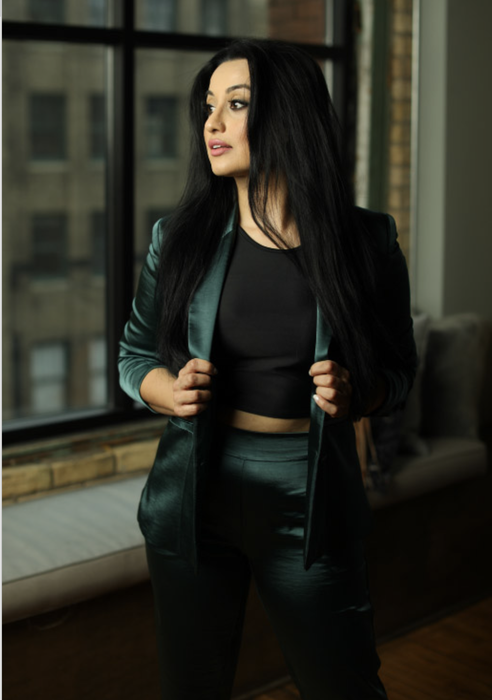 A woman with long black hair standing indoors near a window, wearing a shiny green blazer, a black crop top, and matching green pants.