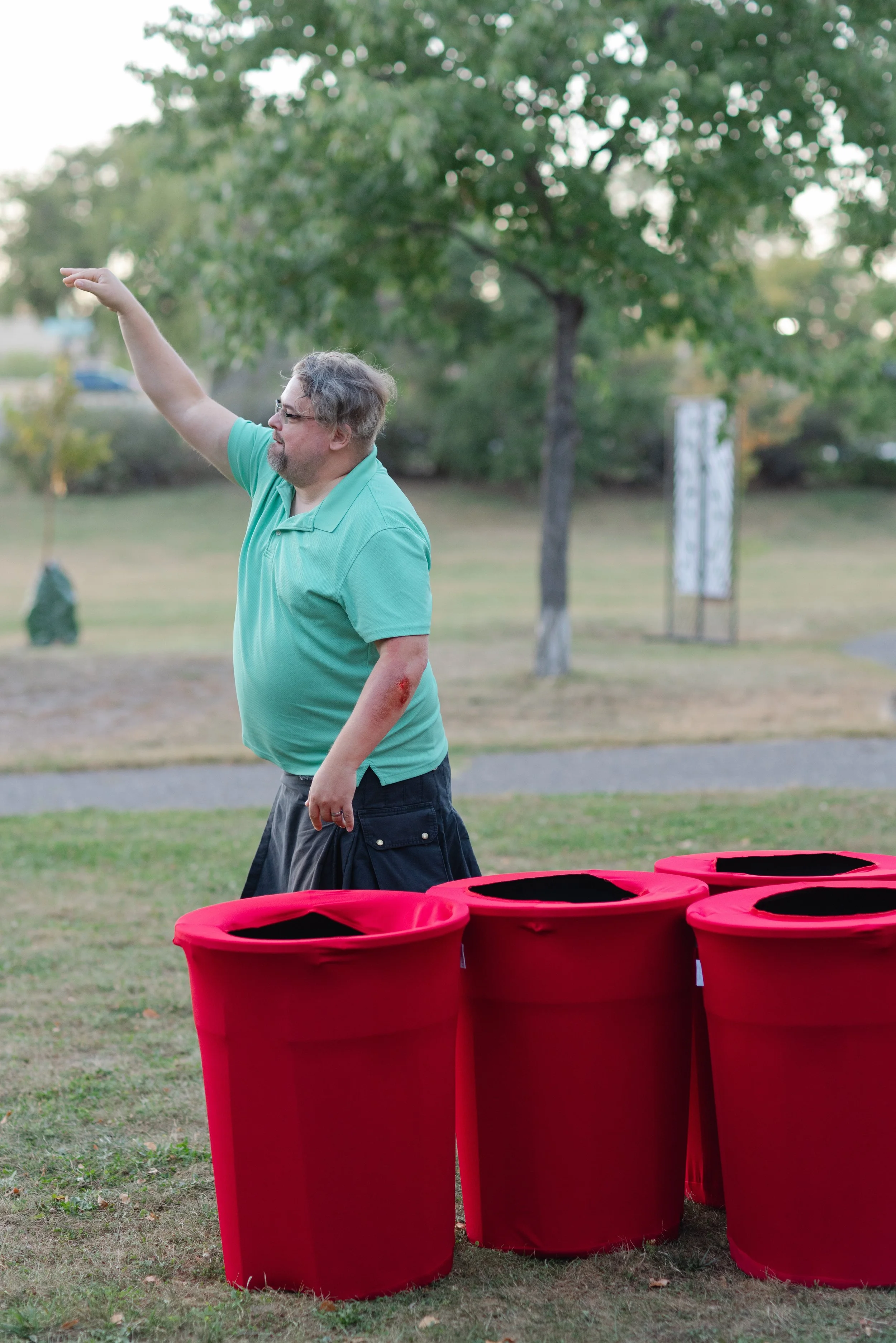 A man in a teal polo shirt with a red bruise or injury on his left arm is standing outdoors in a park, raising his right arm and pointing upward. There are several large pink bins in the foreground, and trees and a walkway in the background.