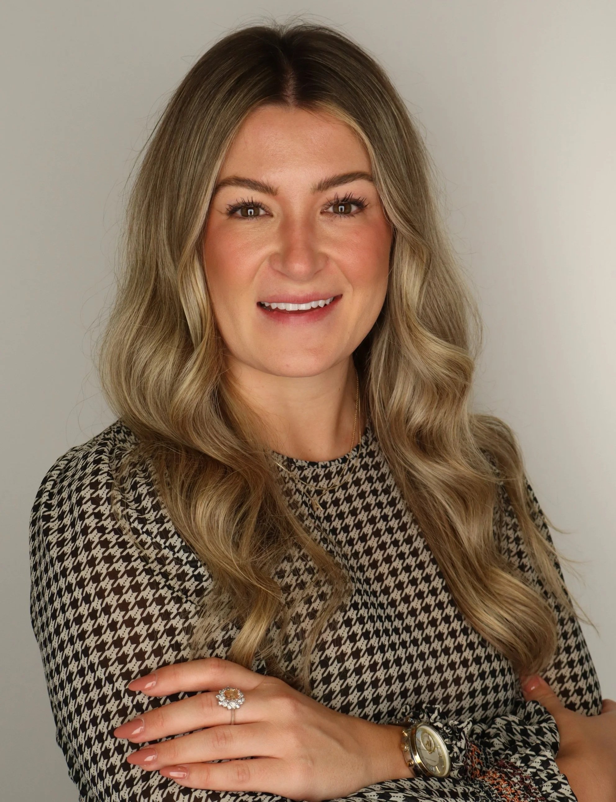 Alyssa Mance A woman with long wavy blonde hair smiling, wearing a patterned top, a watch, and a ring, posing with arms crossed against a plain background.