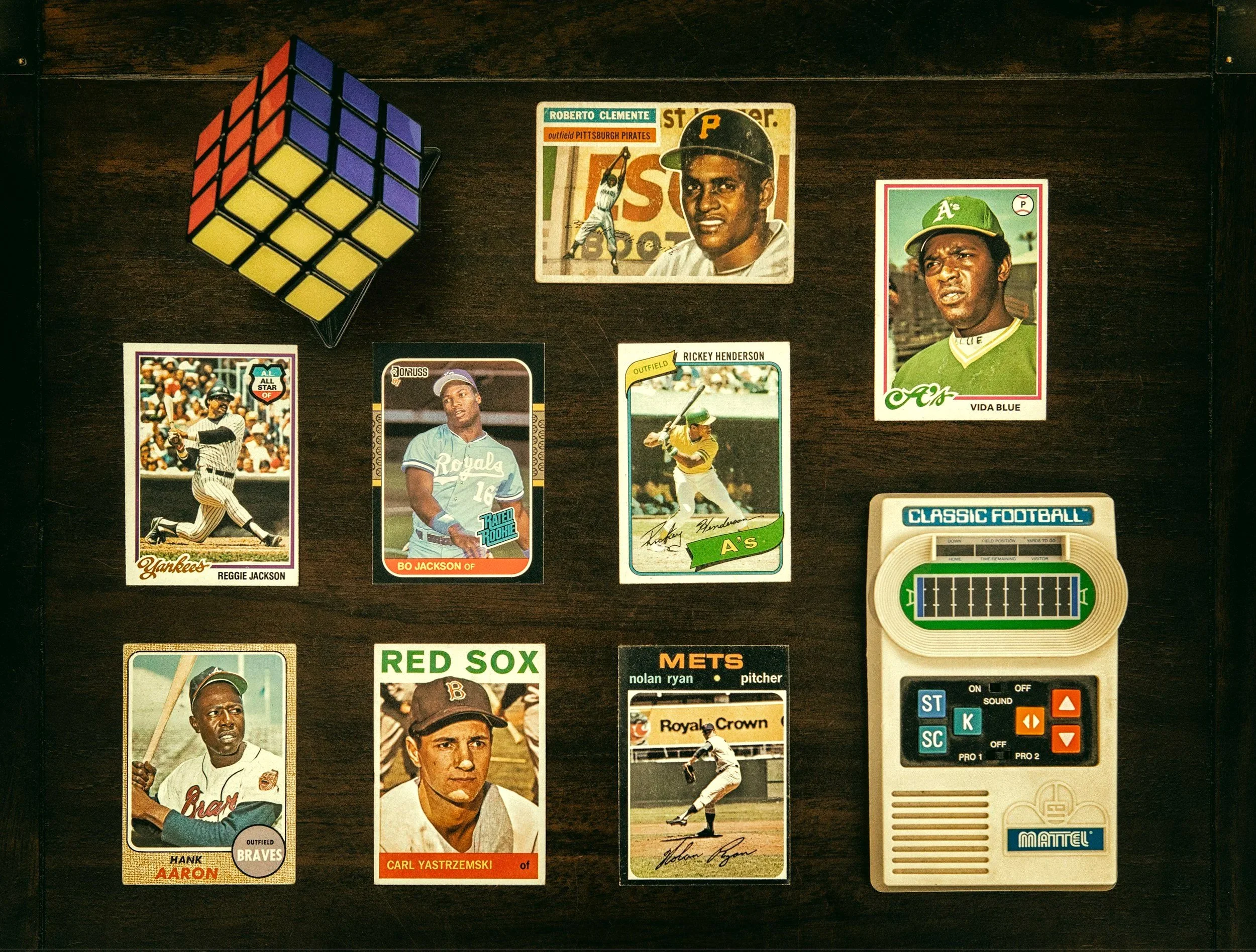 Vintage football and baseball trading cards arranged on a wooden surface, with a 3x3 Rubik's Cube in the top left corner and a classic football game electronic handheld device in the bottom right corner.