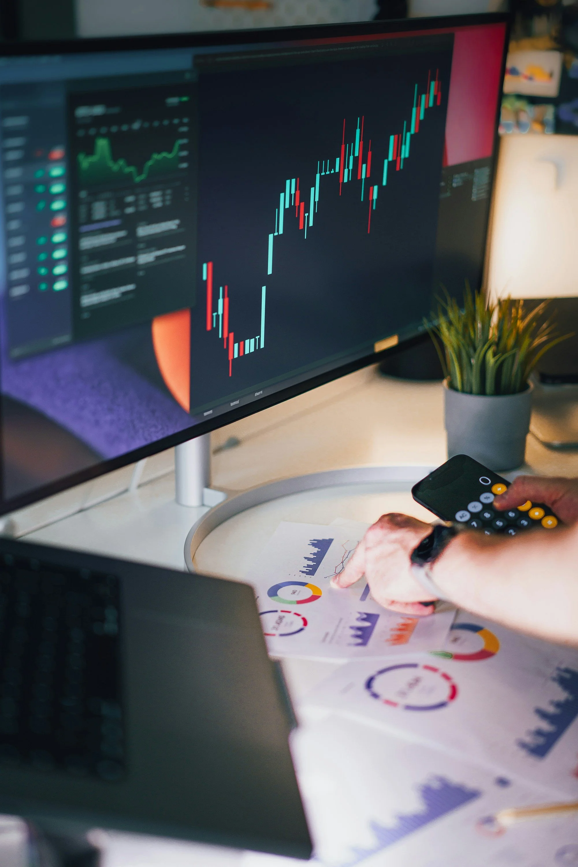 A person analyzing financial data on printed charts and graphs, using a calculator and a computer monitor displaying stock market charts with candlestick patterns and green and red indicators.