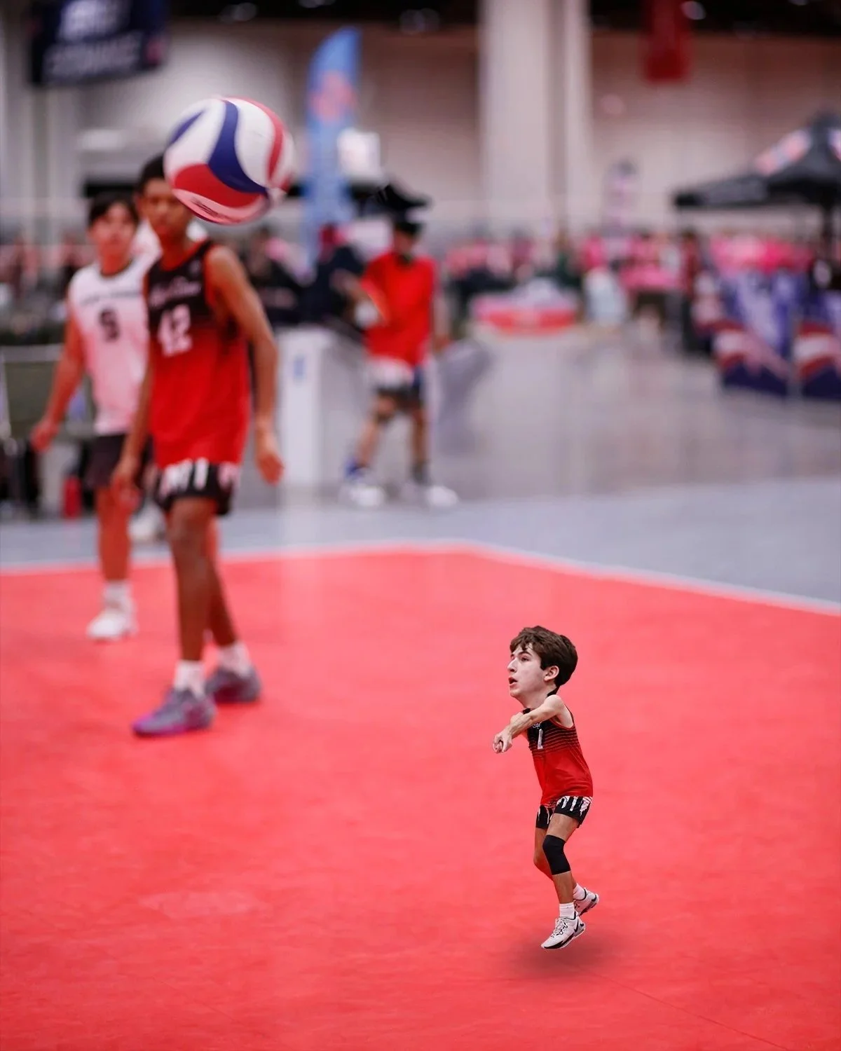 A young boy in a red and black sports uniform playing volleyball on an indoor court, with a volleyball in mid-air near the boy's head, and several other players and people blurred in the background.